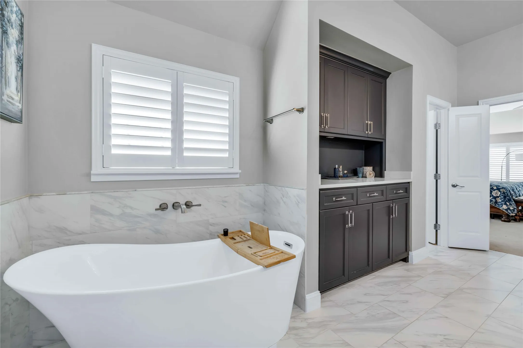 Freestanding soaking tub with wall-mounted fixtures, accented by plantation shutters and a marble-patterned tile wainscoting