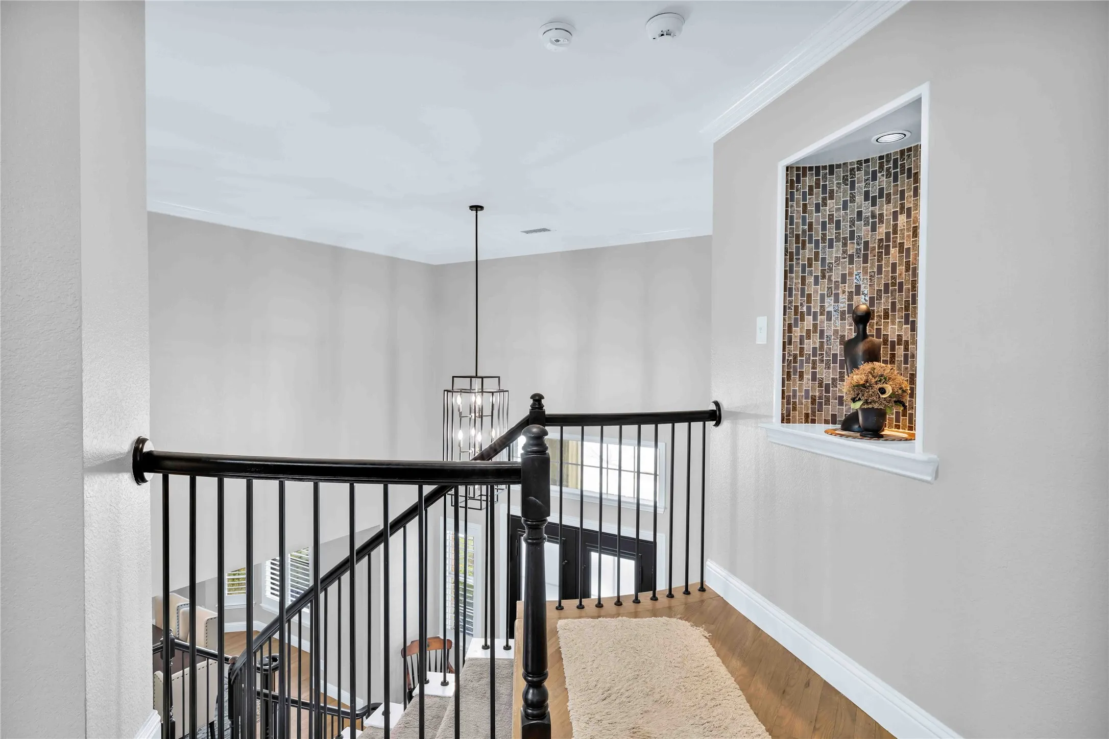 Upper-level hallway featuring wood-finish flooring, a decorative niche with mosaic tile backing, crown molding, and a wrought iron baluster railing