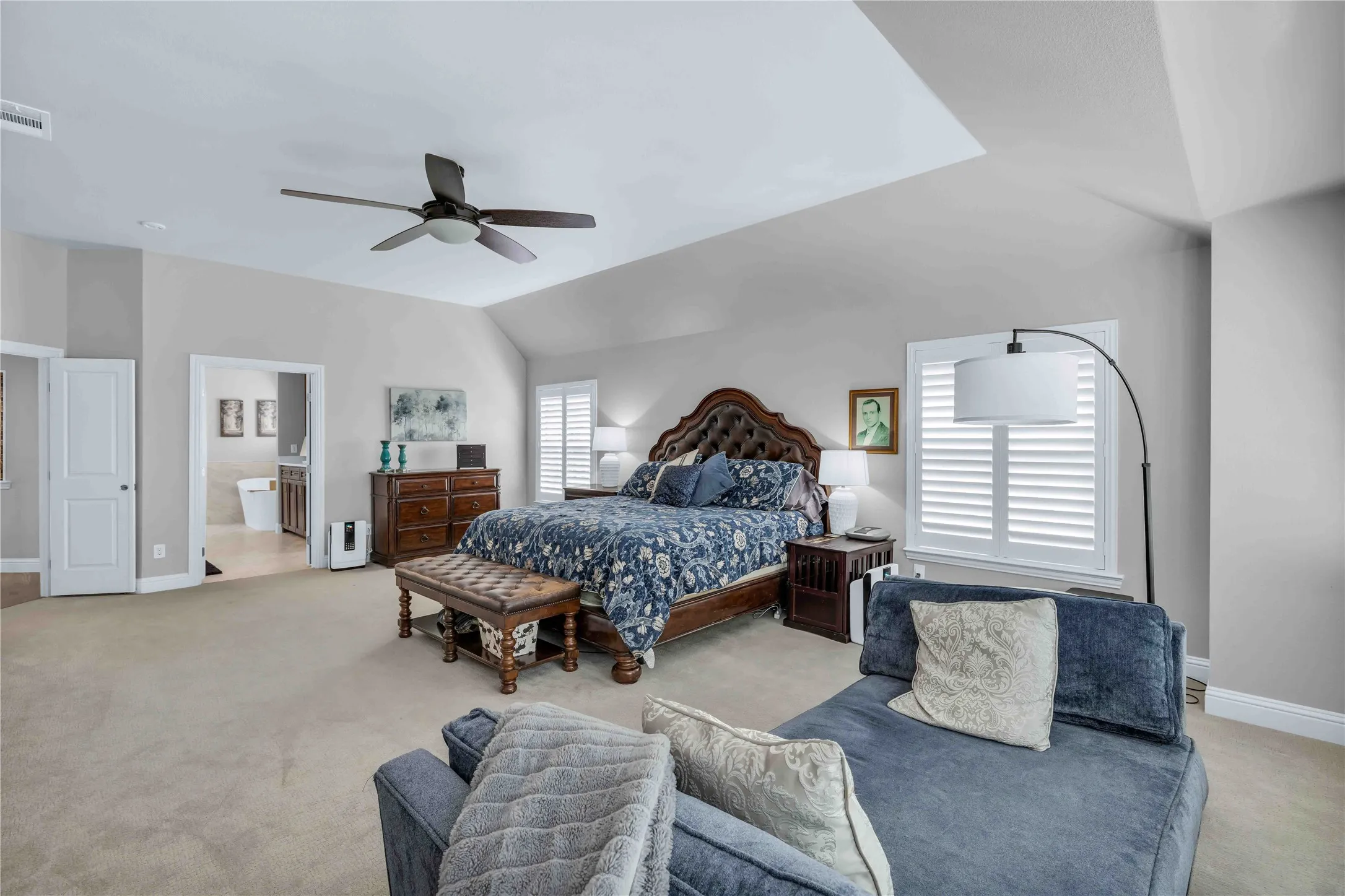 Vaulted ceiling with a ceiling fan, light gray wall paint, plantation shutters, carpet flooring, and an en-suite bathroom entry