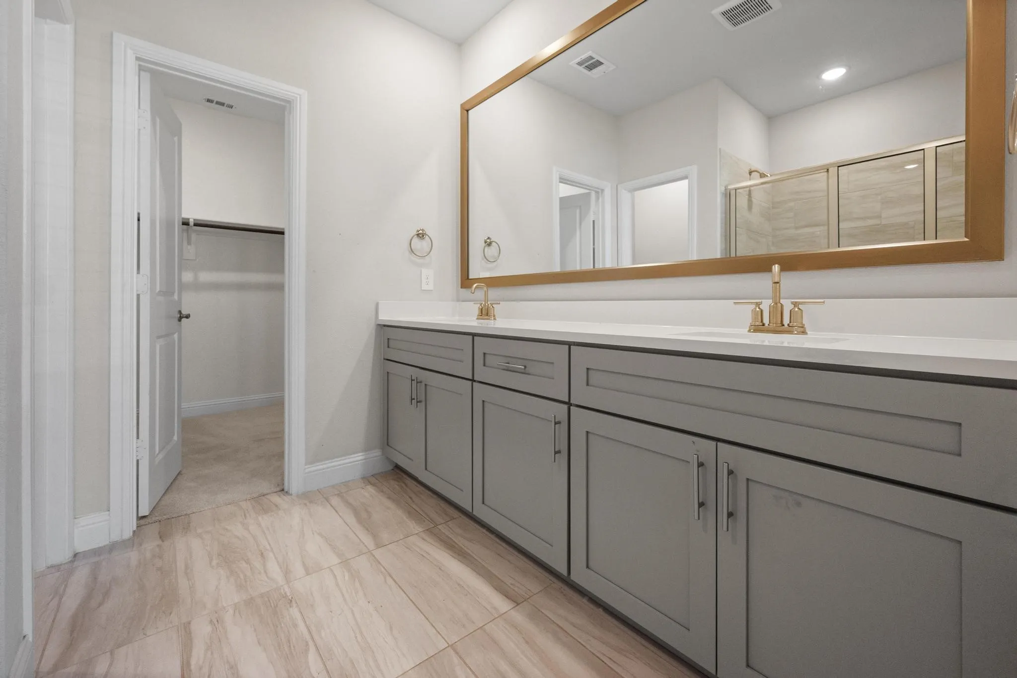 Bathroom featuring dual vanity with gray cabinetry, white countertops, gold-finish fixtures, and a large gold-framed mirror