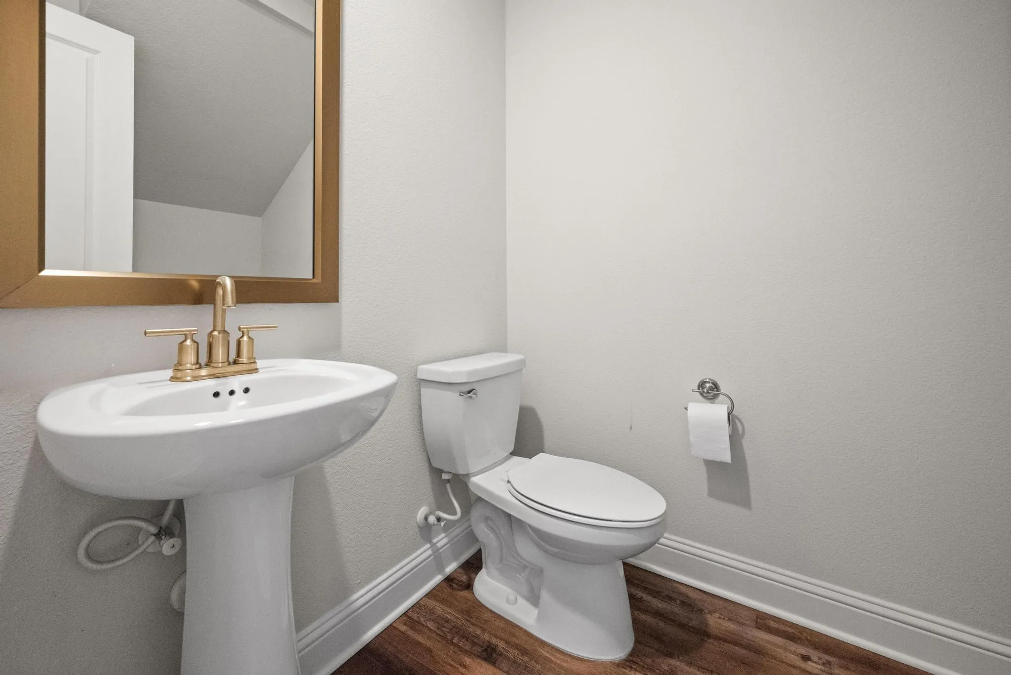 Pedestal sink with gold-tone fixtures, framed mirror, white toilet, and wood-finish flooring