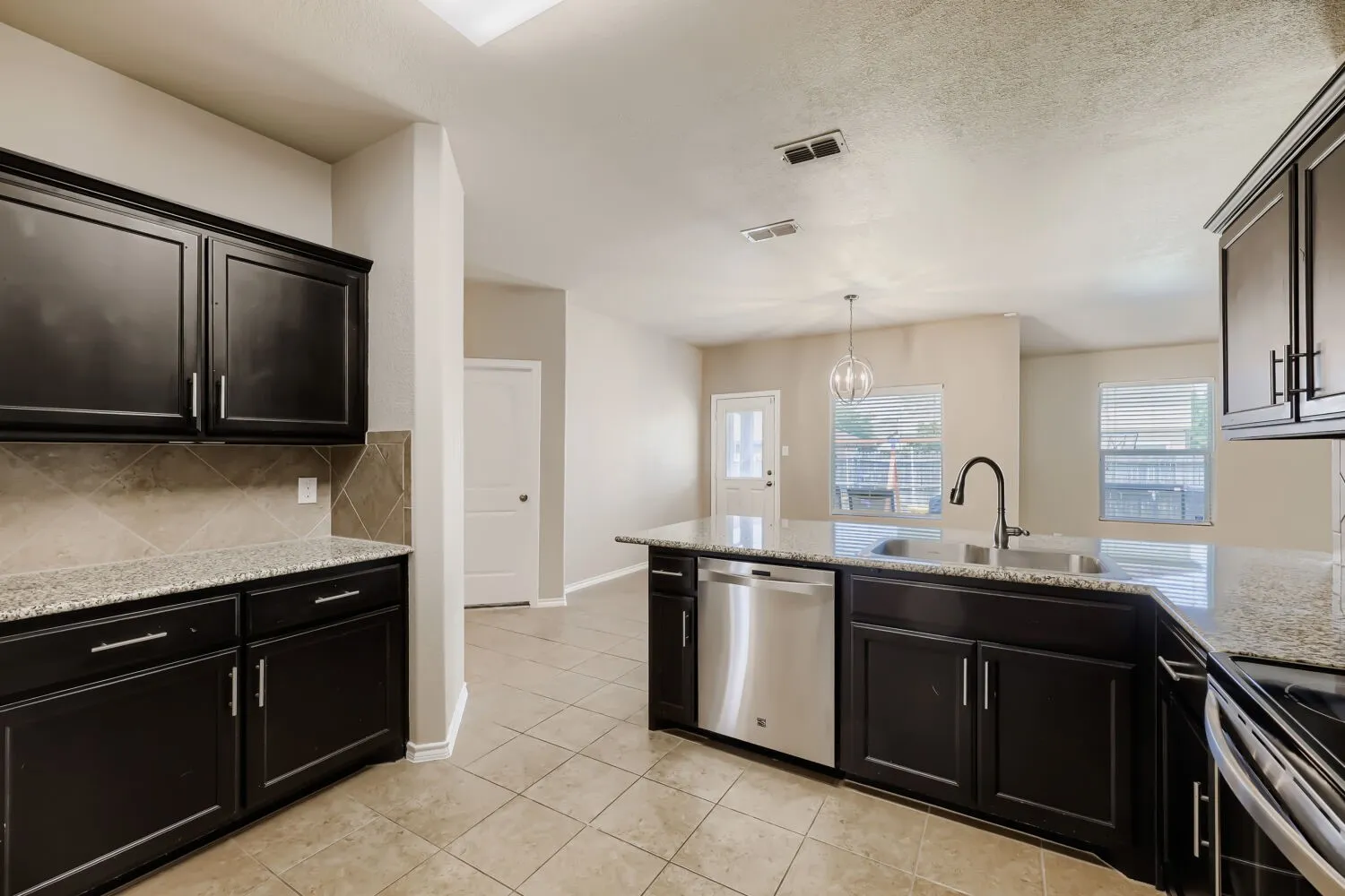 Spacious kitchen featuring black cabinetry with brushed nickel hardware, stainless steel appliances including a double oven and dishwasher, granite countertops with coordinating backsplash, a large center island with prep sink, recessed lighting, and tile flooring throughout