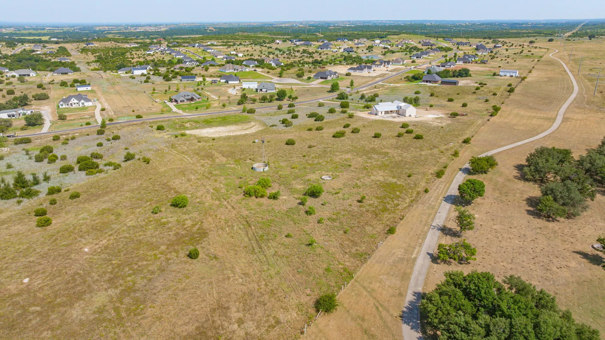 Expansive rural landscape featuring a paved roadway, scattered mature trees, and a distant residential area