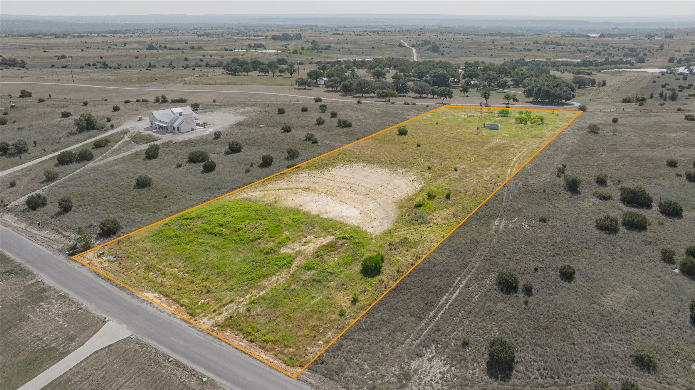 Expansive undeveloped land featuring a partial paved road frontage, varying terrain with areas of green vegetation, and distant trees