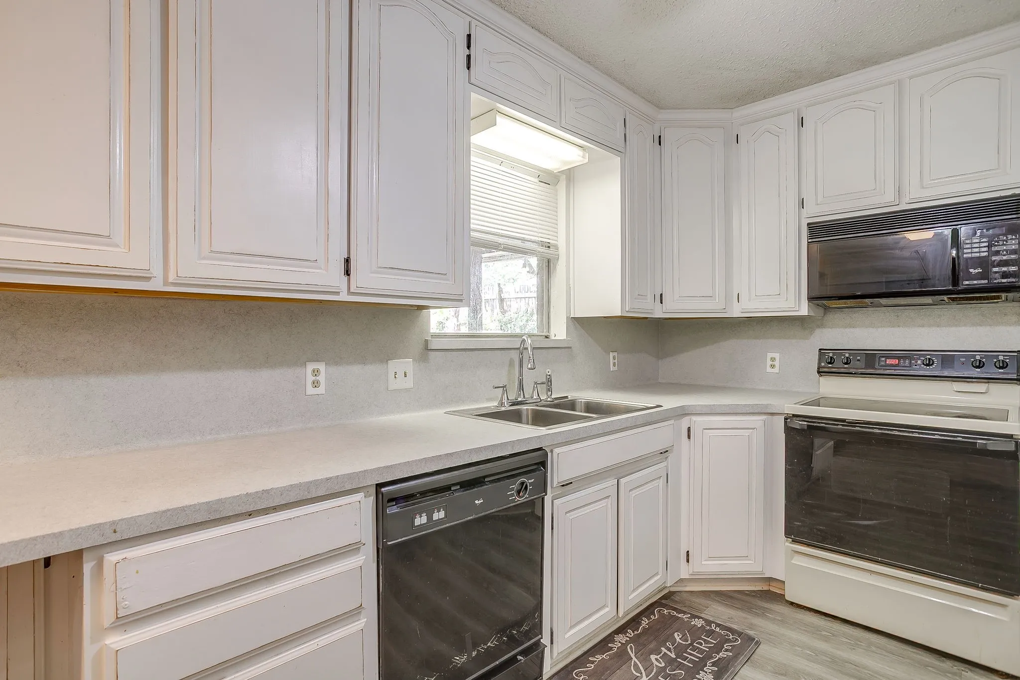 Kitchen featuring white cabinetry, light-toned countertops, a double basin stainless steel sink, black appliances, and wood-finish flooring