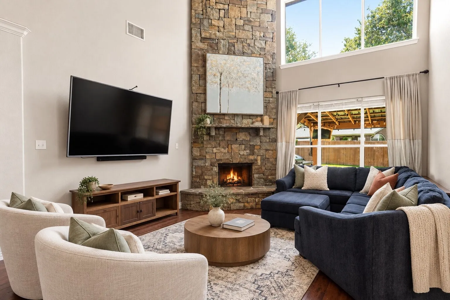 Impressive living room featuring soaring ceilings, a floor-to-ceiling stone fireplace, rich wood flooring, and oversized windows that fill the space with natural light. Photos have been AI enhanced for presentation; no material property features were altered.