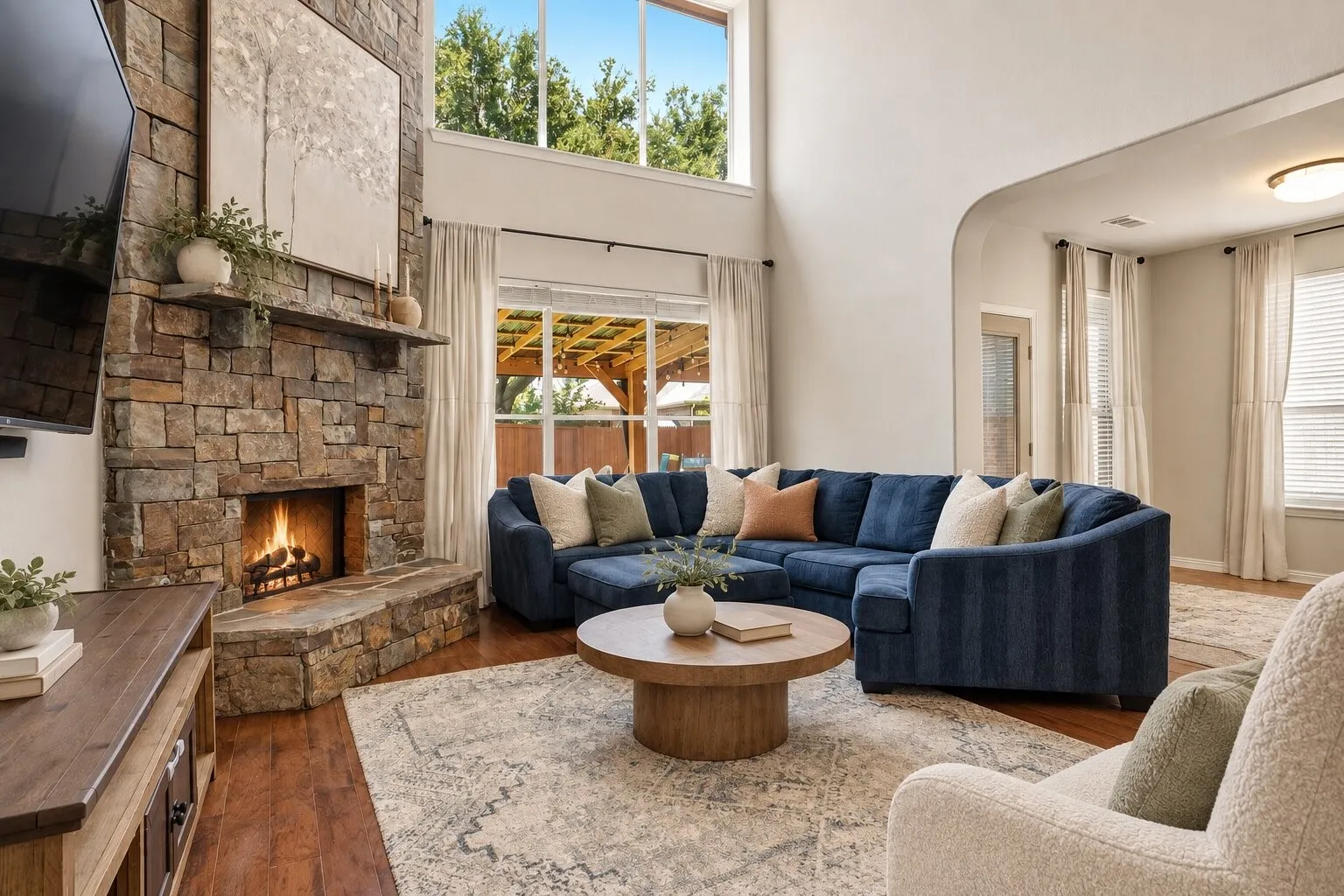 Spacious two-story living room with soaring ceilings, a floor-to-ceiling stone fireplace, rich wood flooring, and large windows overlooking the backyard and covered outdoor living area. Photos have been AI enhanced for presentation; no material property features were altered.