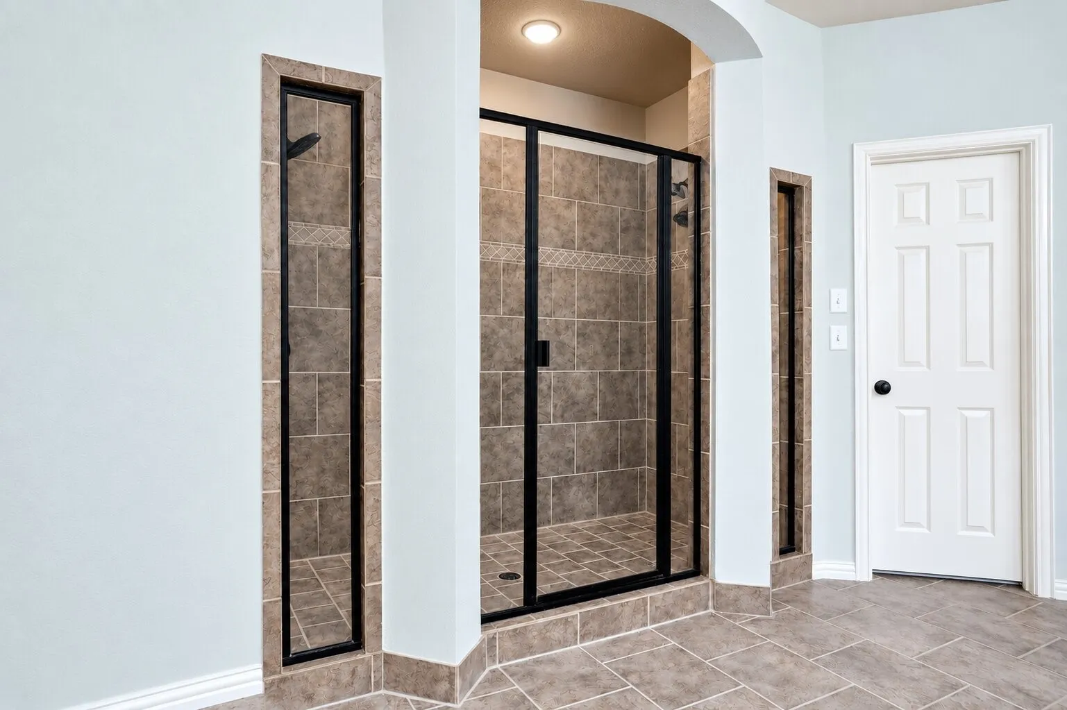 Spacious walk-in shower featuring dual showerheads, a glass enclosure, decorative tilework, and natural light from two narrow windows.