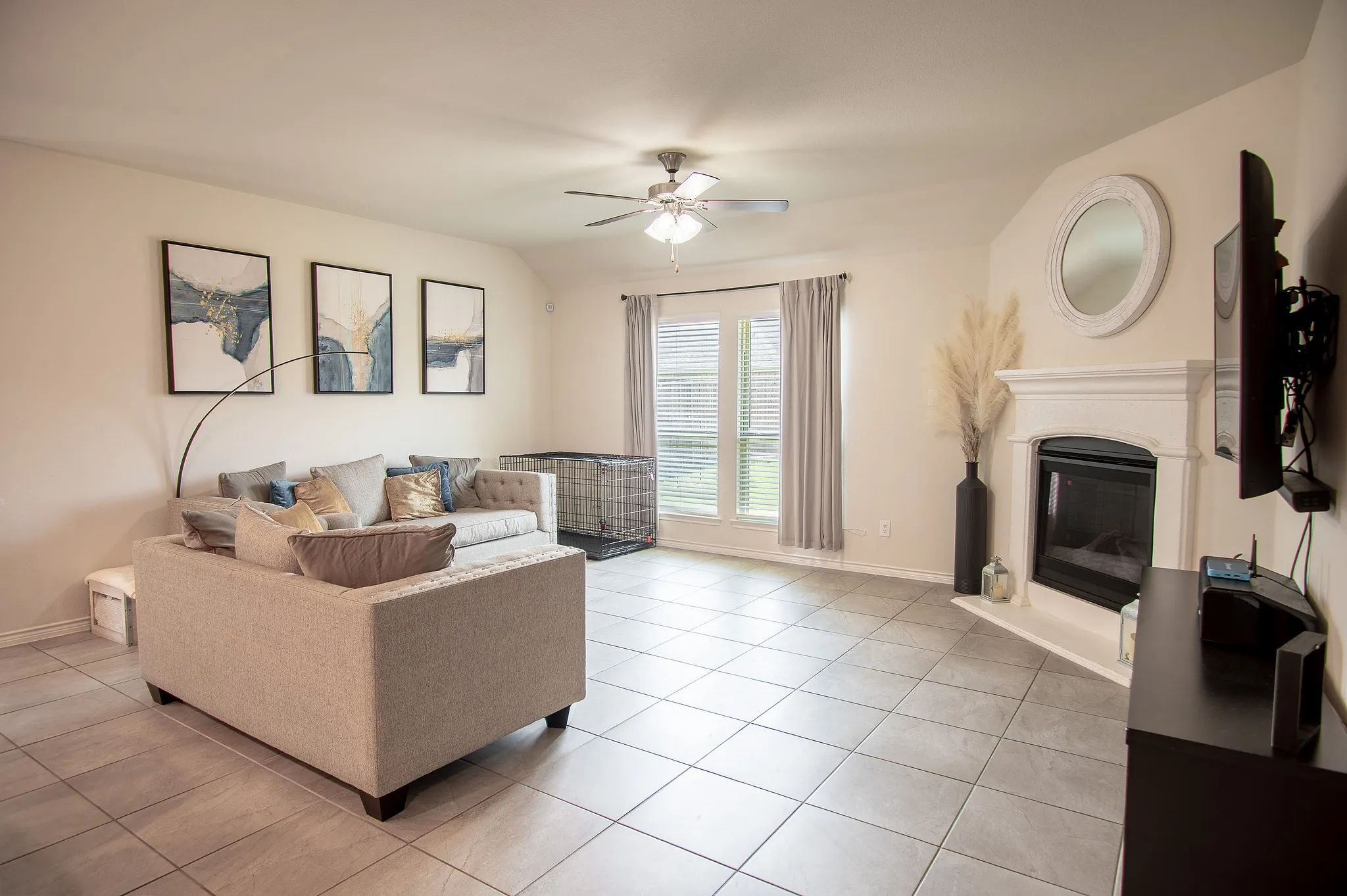 Spacious living area featuring neutral-toned tile flooring, a built-in corner fireplace with a white mantel, and a ceiling fan