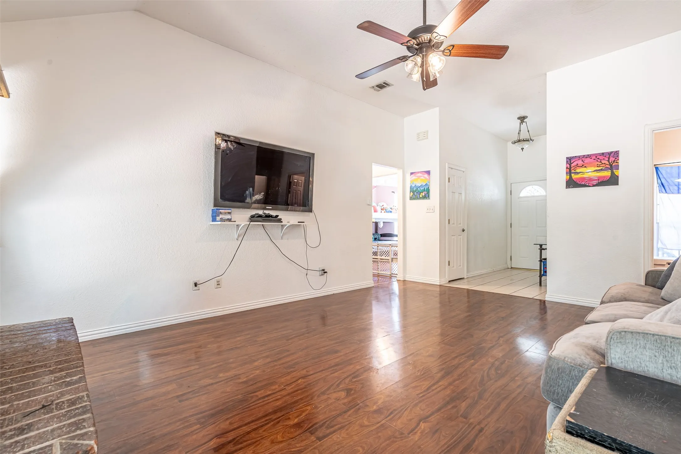 Spacious living area featuring rich wood-finish flooring, ceiling fan with integrated lighting, wall-mounted flat-screen television, and abundant natural light from multiple windows