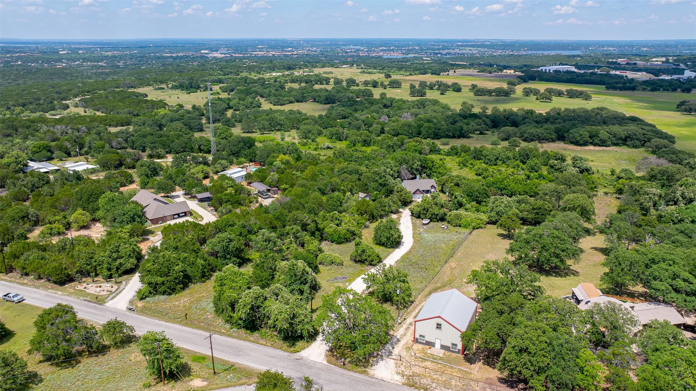 Scenic aerial perspective showcasing this rural estate nestled among mature oak and pine trees, with a winding private driveway, multiple outbuildings, and expansive acreage bordered by open pastureland and rolling countryside