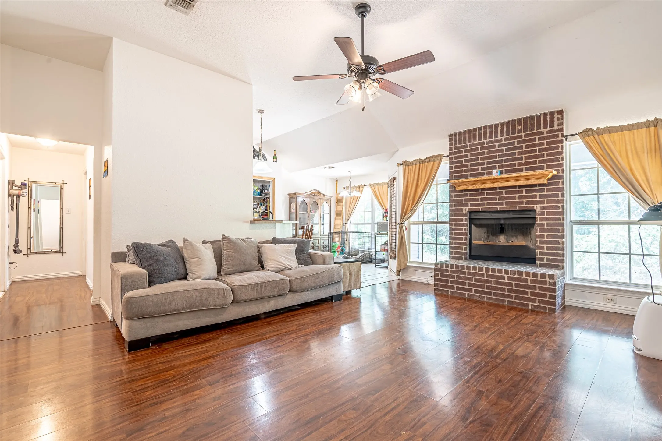 Spacious living area featuring wood-finish flooring, a brick fireplace with a mantel, vaulted ceilings, a ceiling fan, and large windows