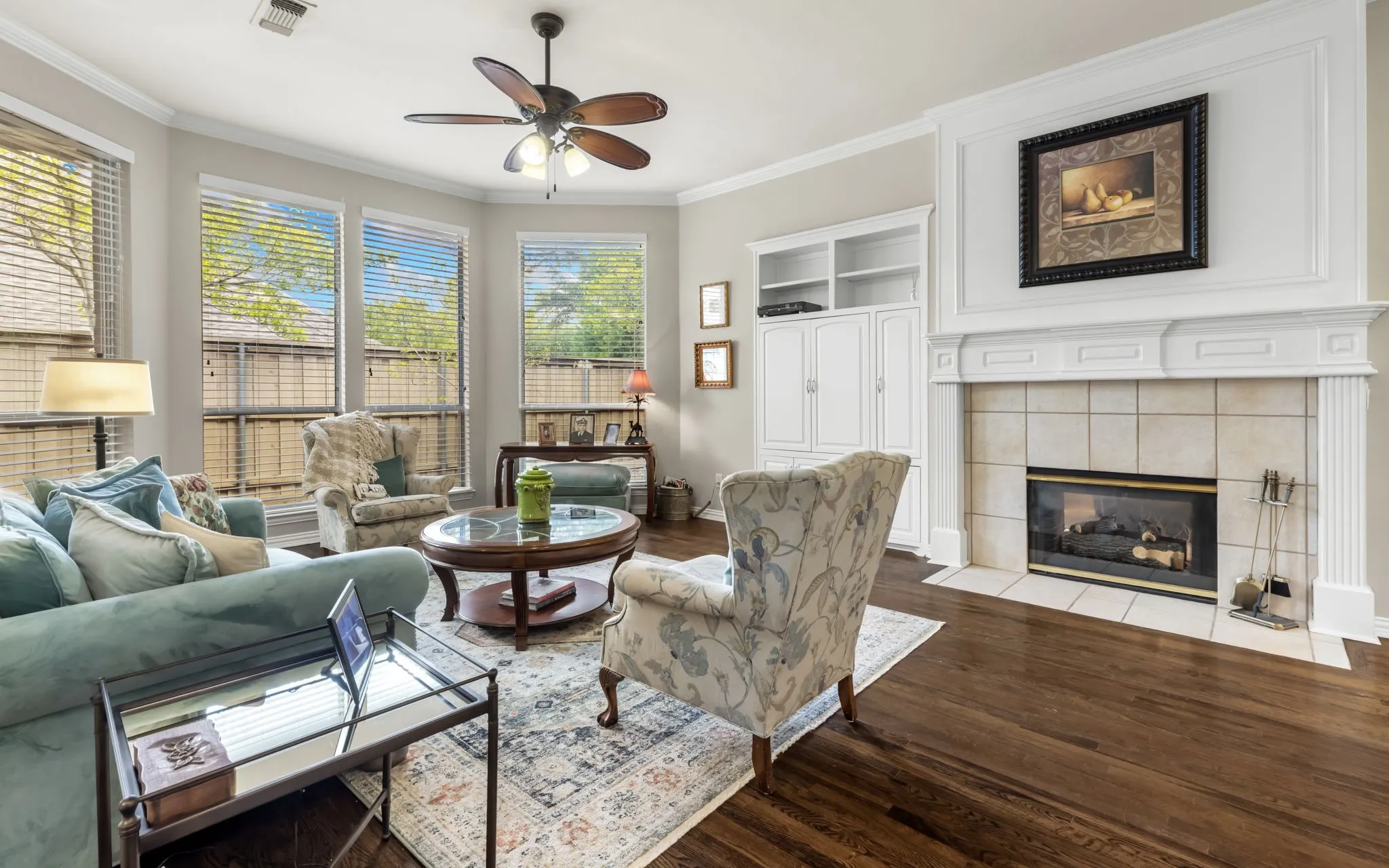 Spacious living area featuring wood-finish flooring, a tiled fireplace with an ornate mantel, built-in cabinetry, large windows, and a ceiling fan