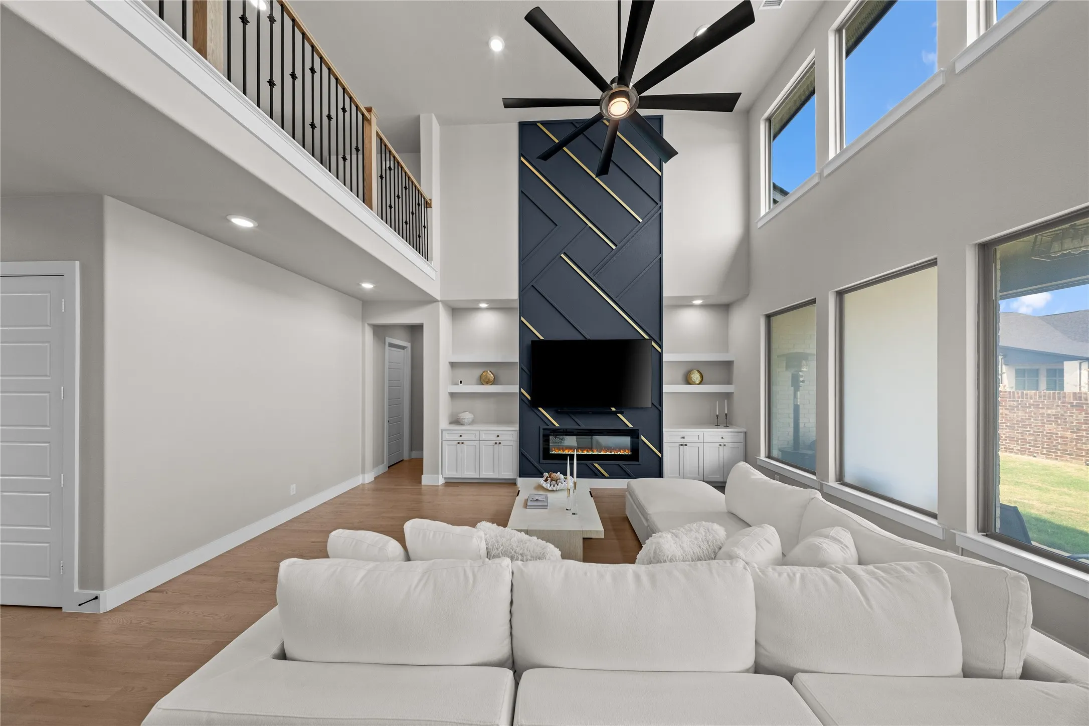 Two-story ceiling, wood-finish flooring, custom accent wall with integrated linear fireplace, built-in shelving with cabinetry, and an upper-level balcony with metal spindle railing