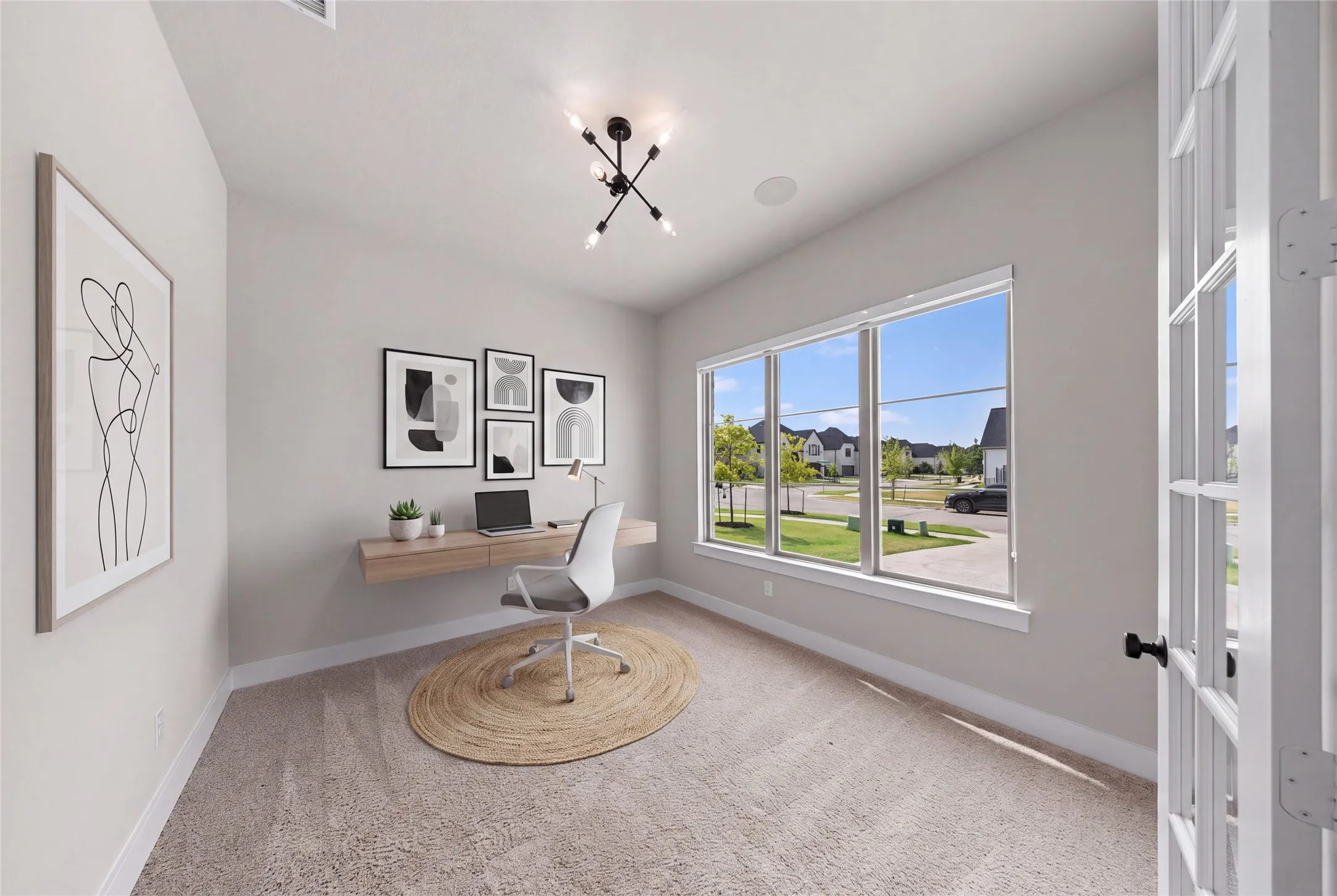 Bright home office featuring expansive multi-pane windows framing natural light and outdoor scenery, light neutral carpeting, a floating desk with integrated storage, modern ceiling fixture, and curated wall art