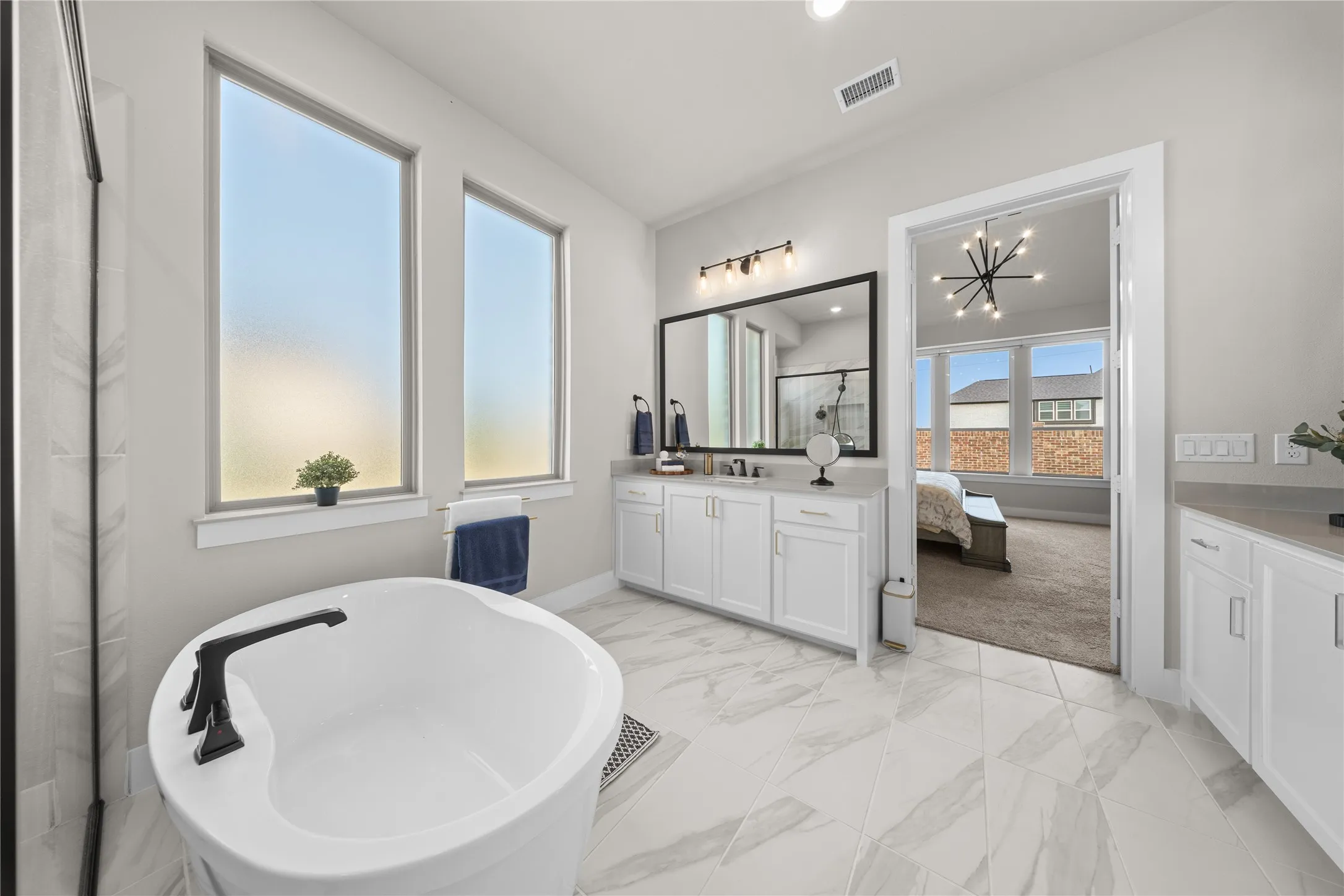 Spa-inspired primary bathroom featuring a freestanding soaking tub, dual-sink vanity with white cabinetry, marble-finish flooring, and large windows with natural light