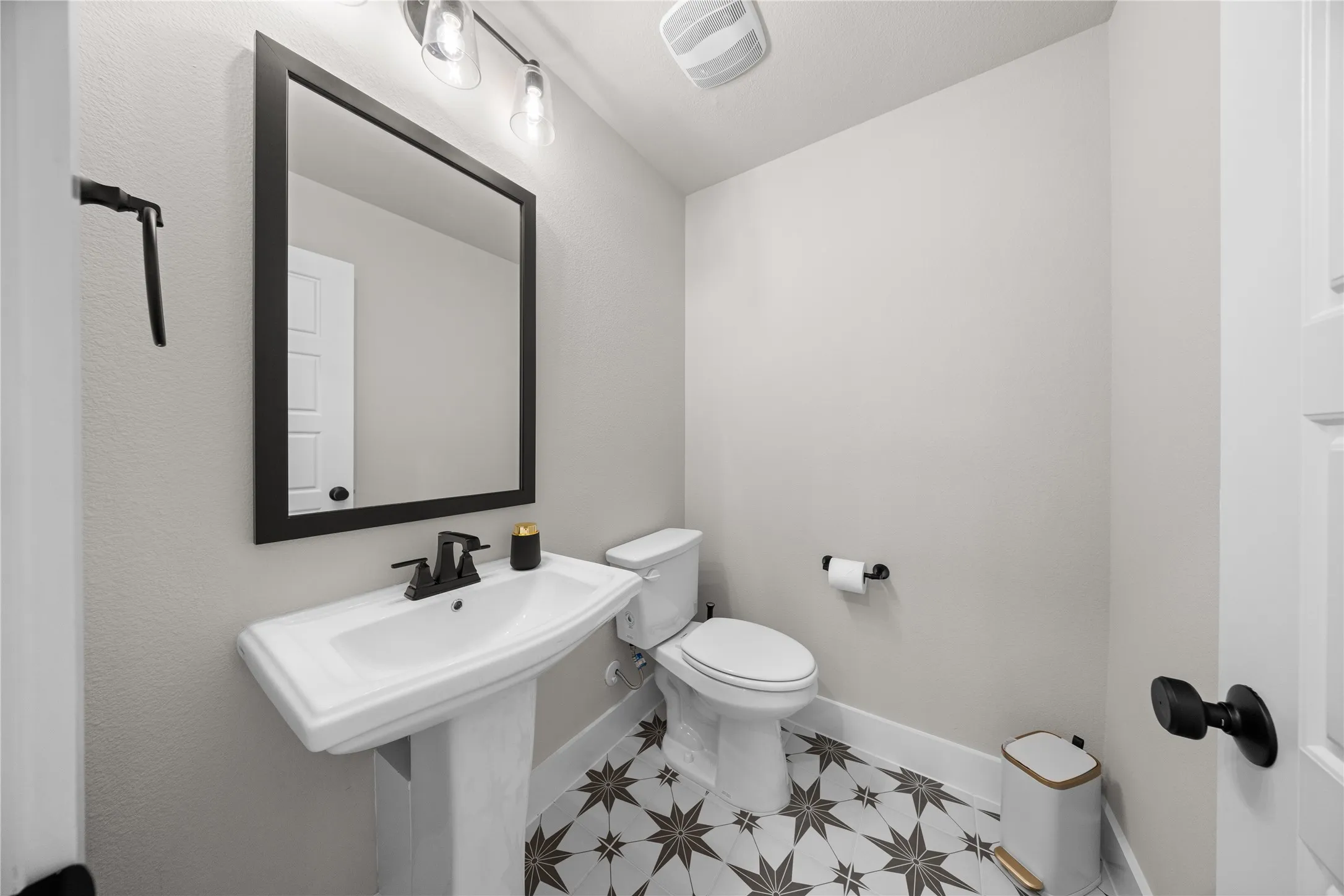 Powder room featuring a pedestal sink with chrome faucet, framed mirror with matte black trim, white ceramic toilet, and patterned tile flooring