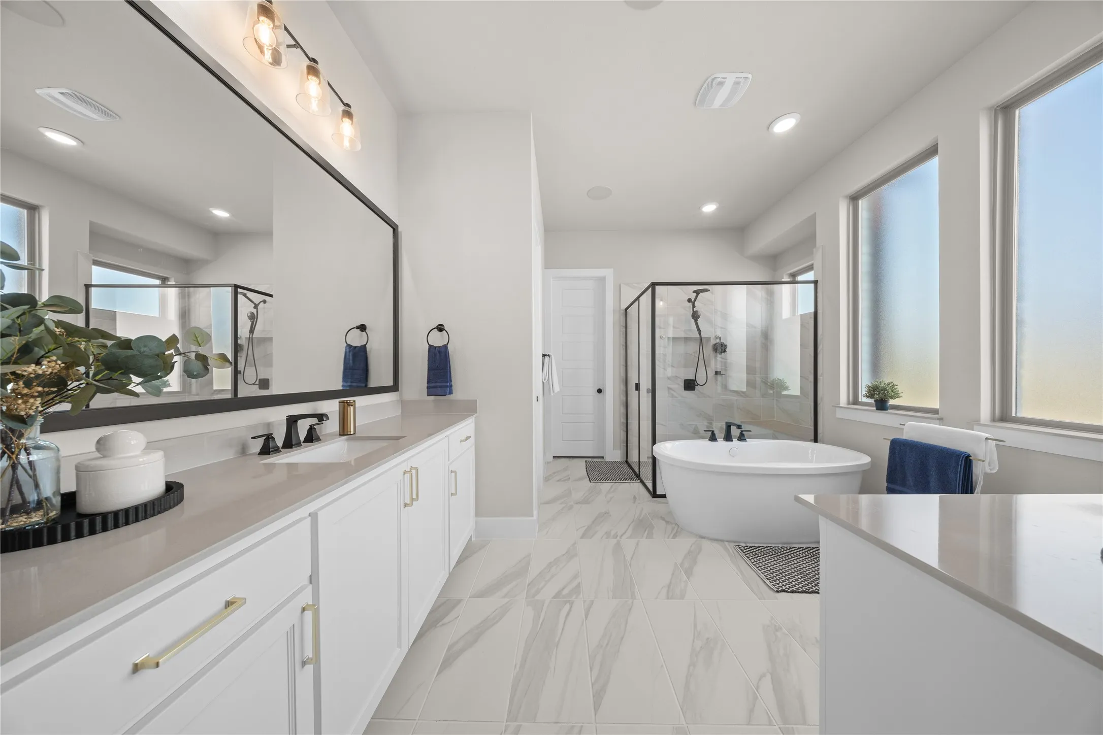 Spa-inspired primary bathroom featuring dual vanities with white cabinetry and quartz countertops, a freestanding soaking tub, walk-in shower with frameless glass enclosure, wide-plank wood-finish flooring, and abundant natural light from expansive windows