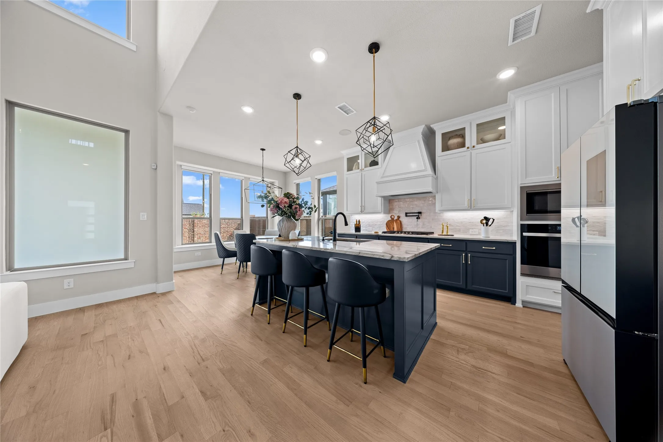 Contemporary kitchen and dining area featuring white cabinetry with integrated appliances, a large waterfall-edge island with seating for multiple guests, wide-plank wood-finish flooring throughout, pendant lighting with geometric fixtures, and abundant natural light from expansive windows and skylights