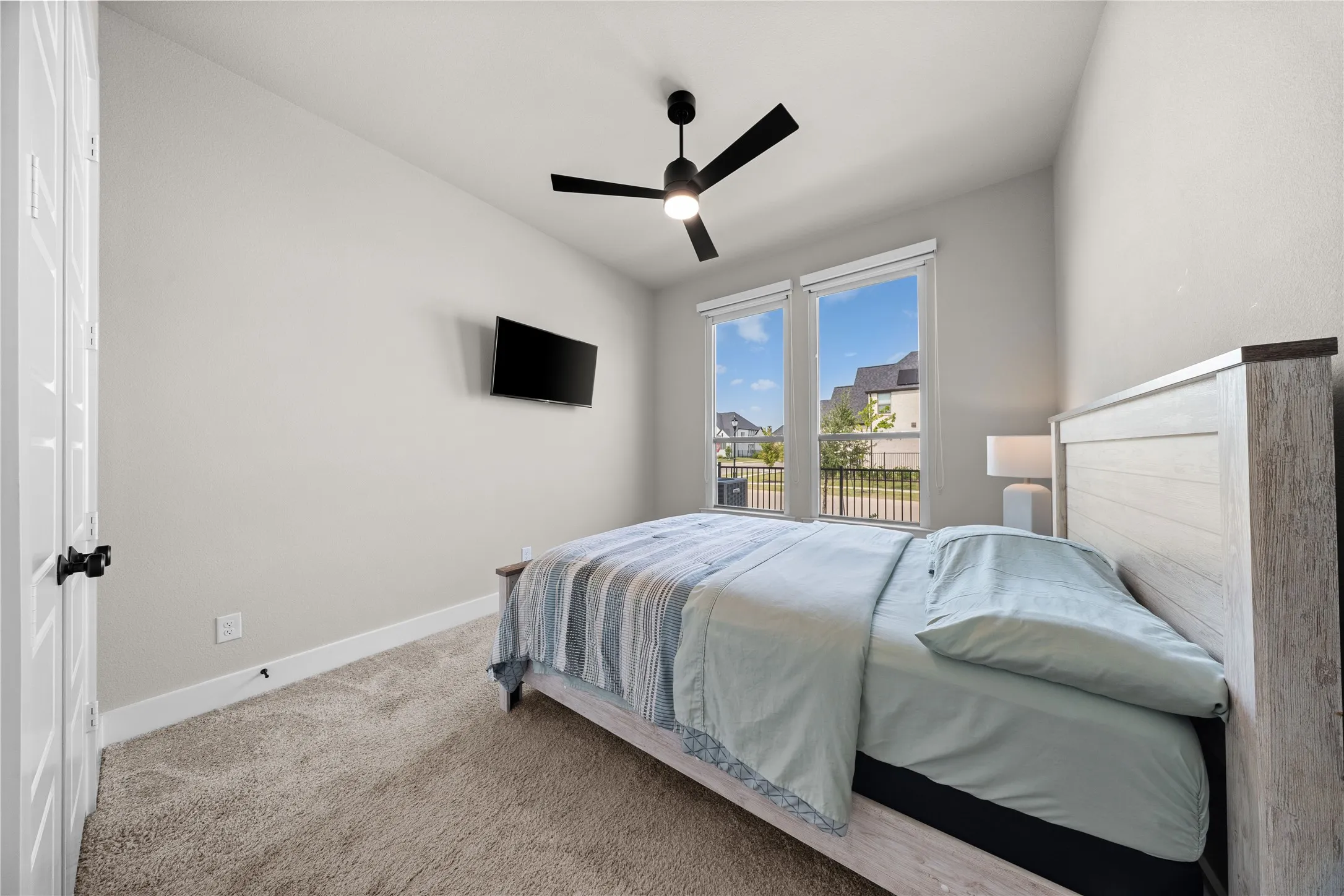 Bright secondary bedroom showcasing carpet flooring, a ceiling fan with black finish, wall-mounted flat-screen television, and glass sliding doors opening to a private balcony with scenic outdoor vistas