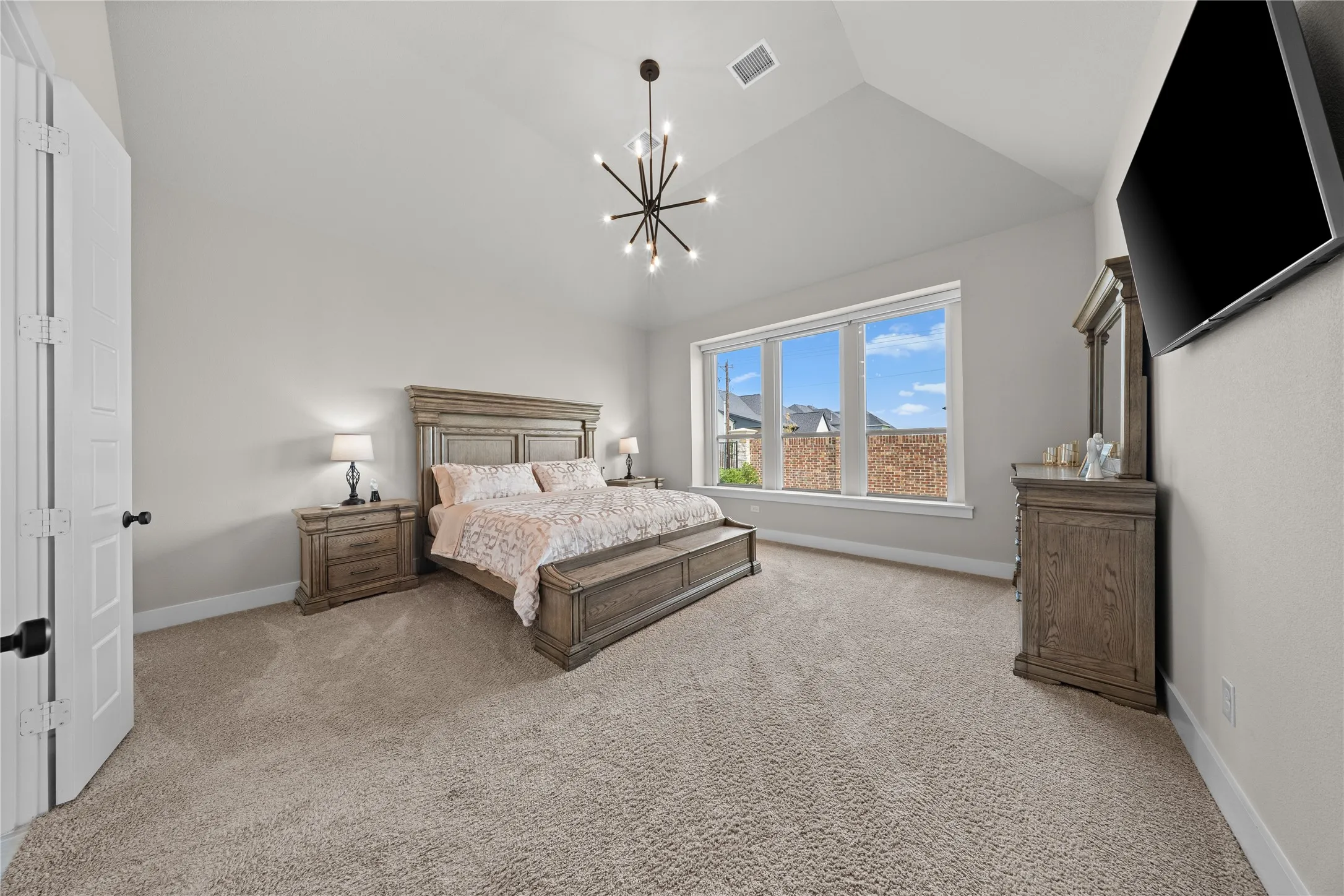 Spacious primary bedroom featuring vaulted ceilings, large multi-pane windows with natural light, neutral carpeting throughout, wood-finish furnishings, and a modern sputnik-style pendant light fixture