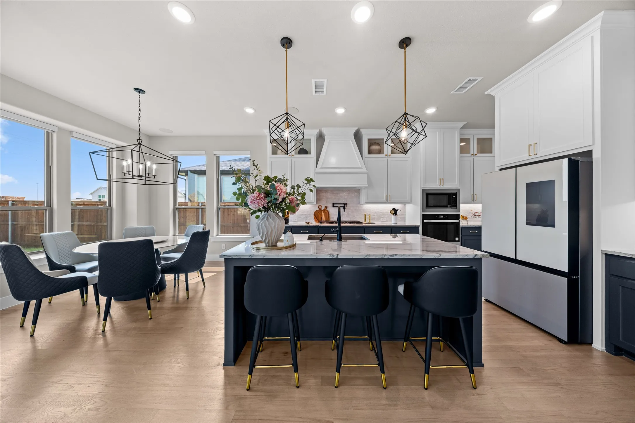 Open-concept kitchen and dining area featuring a large center island with waterfall countertop and seating for multiple guests, stainless steel appliances including a built-in wall oven and range hood, white cabinetry with ample storage, wood-finish flooring throughout, and abundant natural light from expansive windows and glass doors