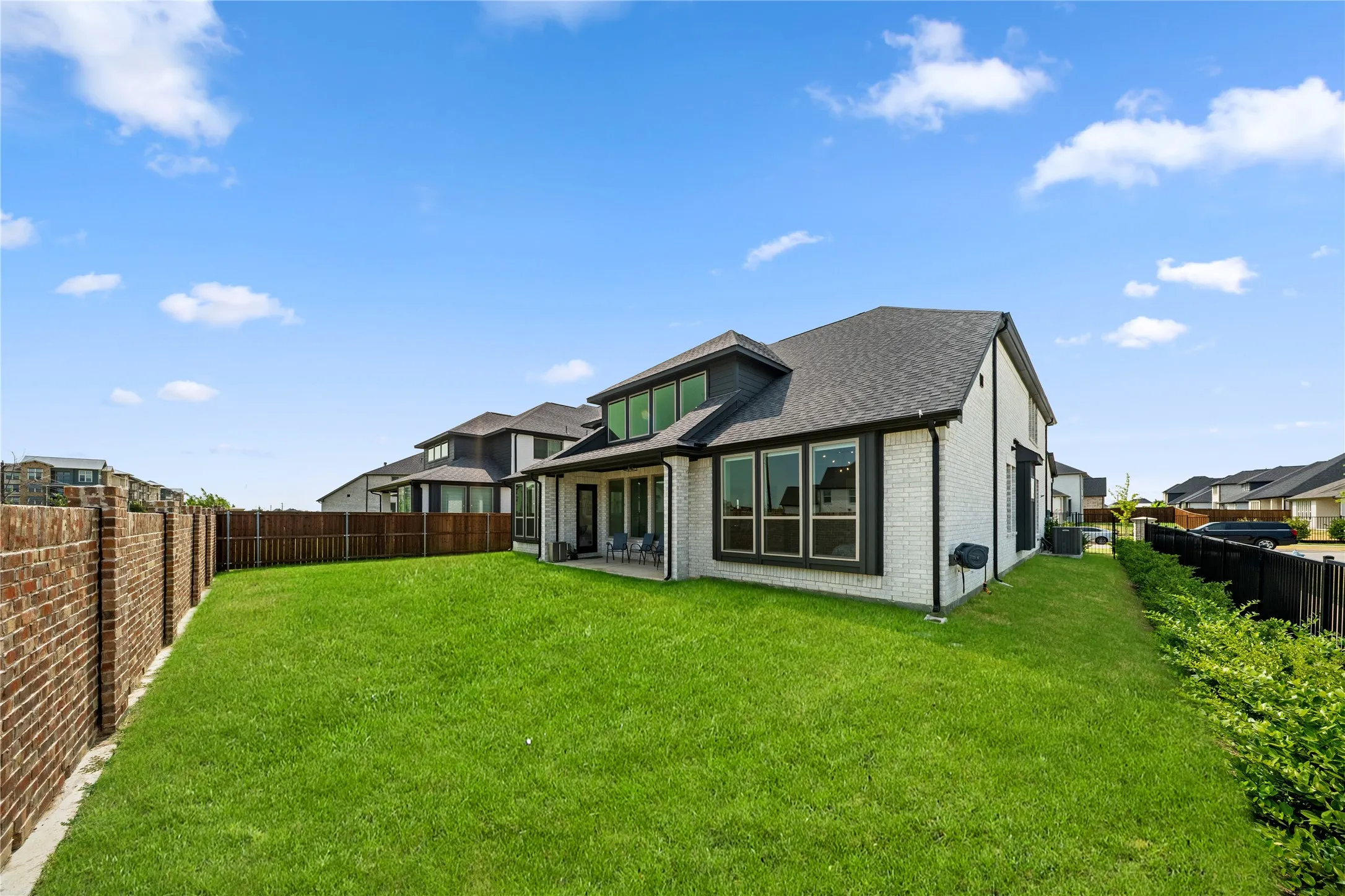 Expansive backyard featuring a manicured lawn bordered by wood fencing, with the residence showcasing a modern architectural design, multiple large windows, and a peaked roofline with dormer detailing