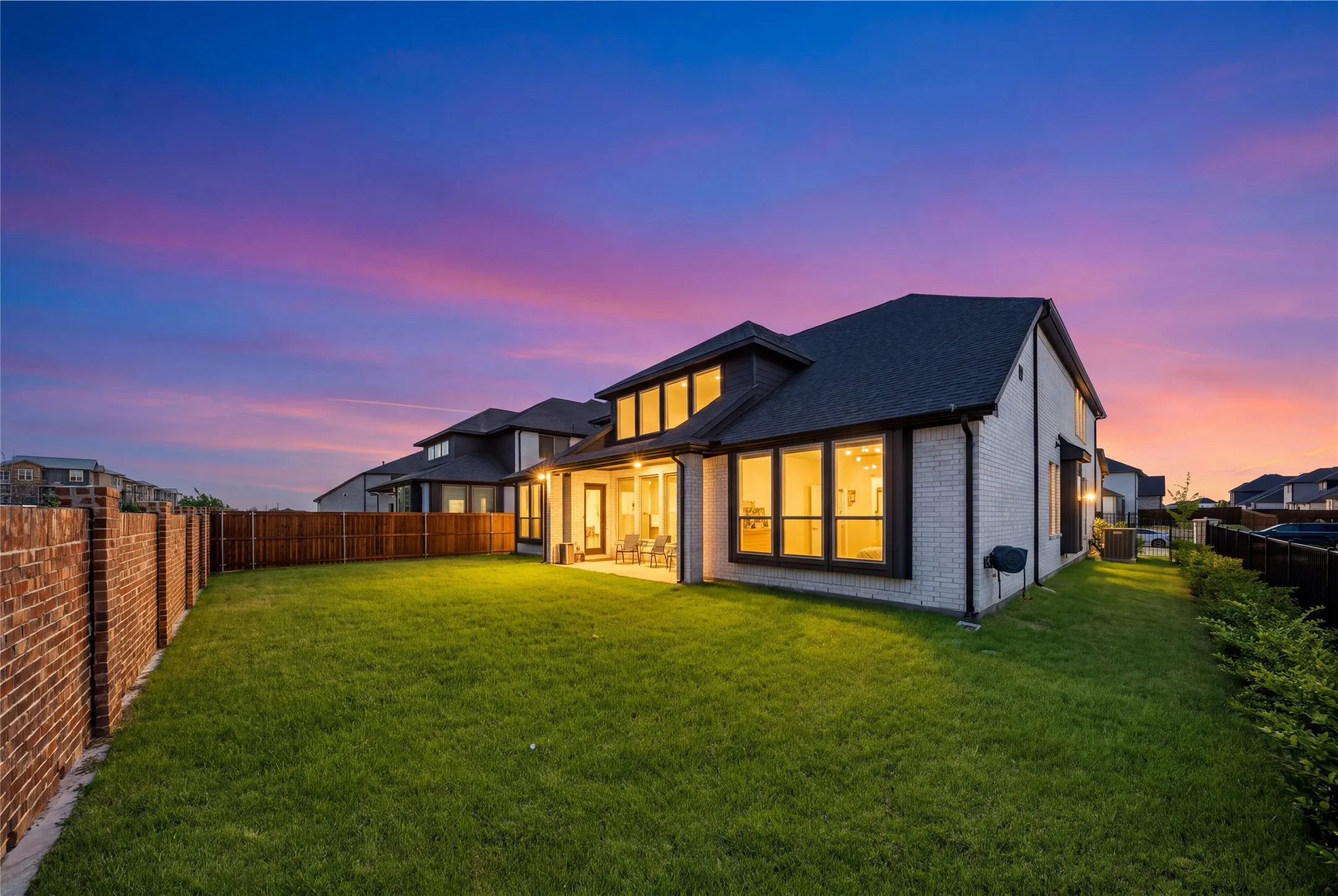 Modern home with illuminated interior spaces, featuring large multi-pane windows, a pitched roof with dormers, mixed exterior finishes including brick and light-colored cladding, and an expansive manicured lawn with defined landscaping borders