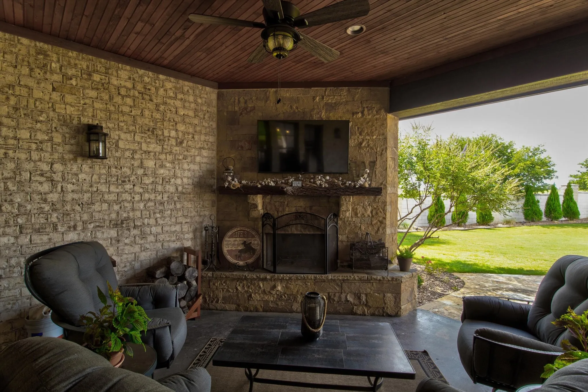 Covered outdoor patio featuring a stone fireplace, wood-plank ceiling with a ceiling fan, light-toned brick wall, and a concrete patio surface