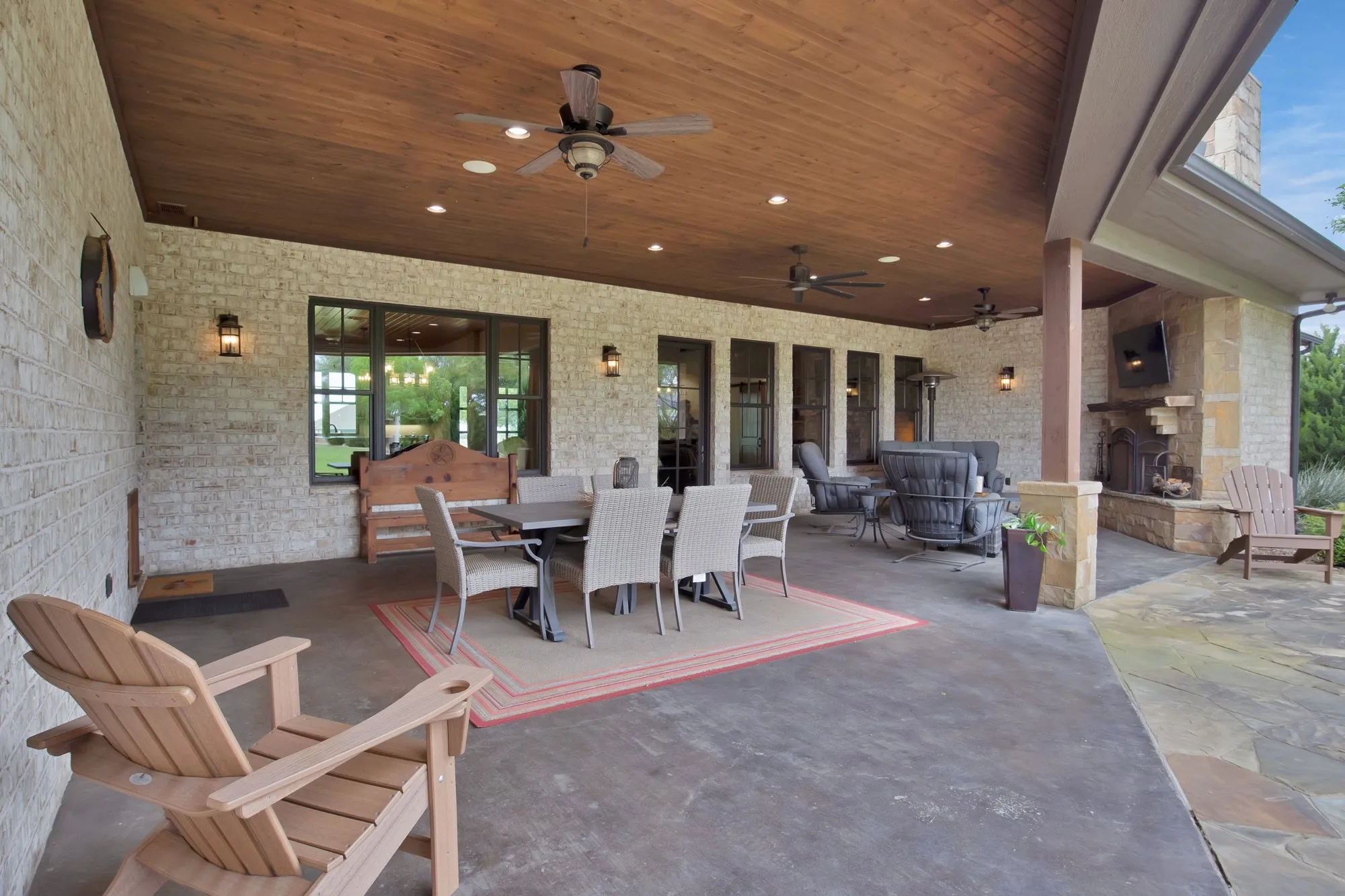 Expansive covered patio featuring a wood-panel ceiling with recessed lighting and ceiling fans, a stone-clad fireplace, and an exterior brick wall