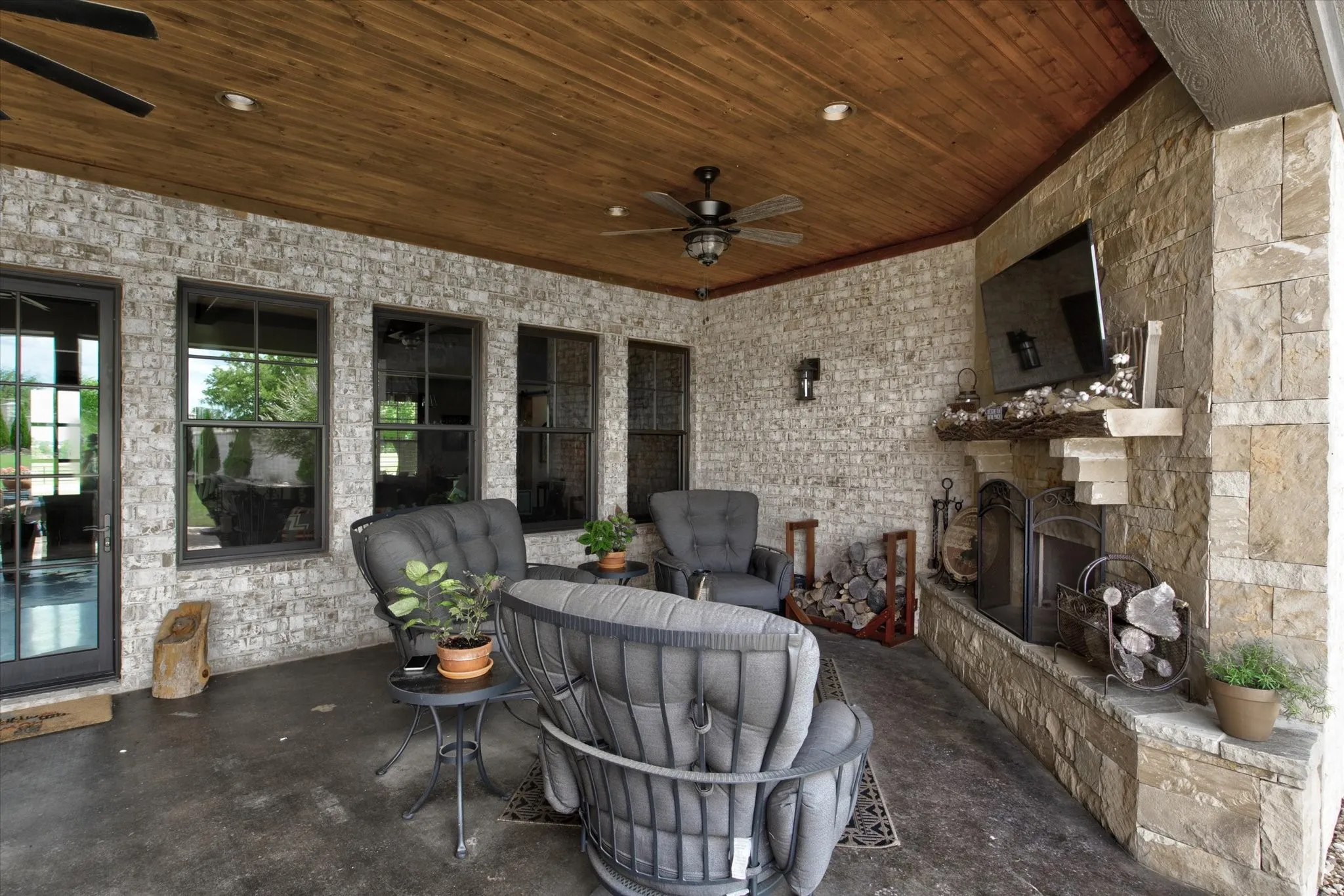 Expansive covered patio featuring multiple French doors, a wood-finish ceiling with ceiling fan, stone fireplace with hearth and mantel, dark concrete flooring, and built-in seating areas with upholstered chairs