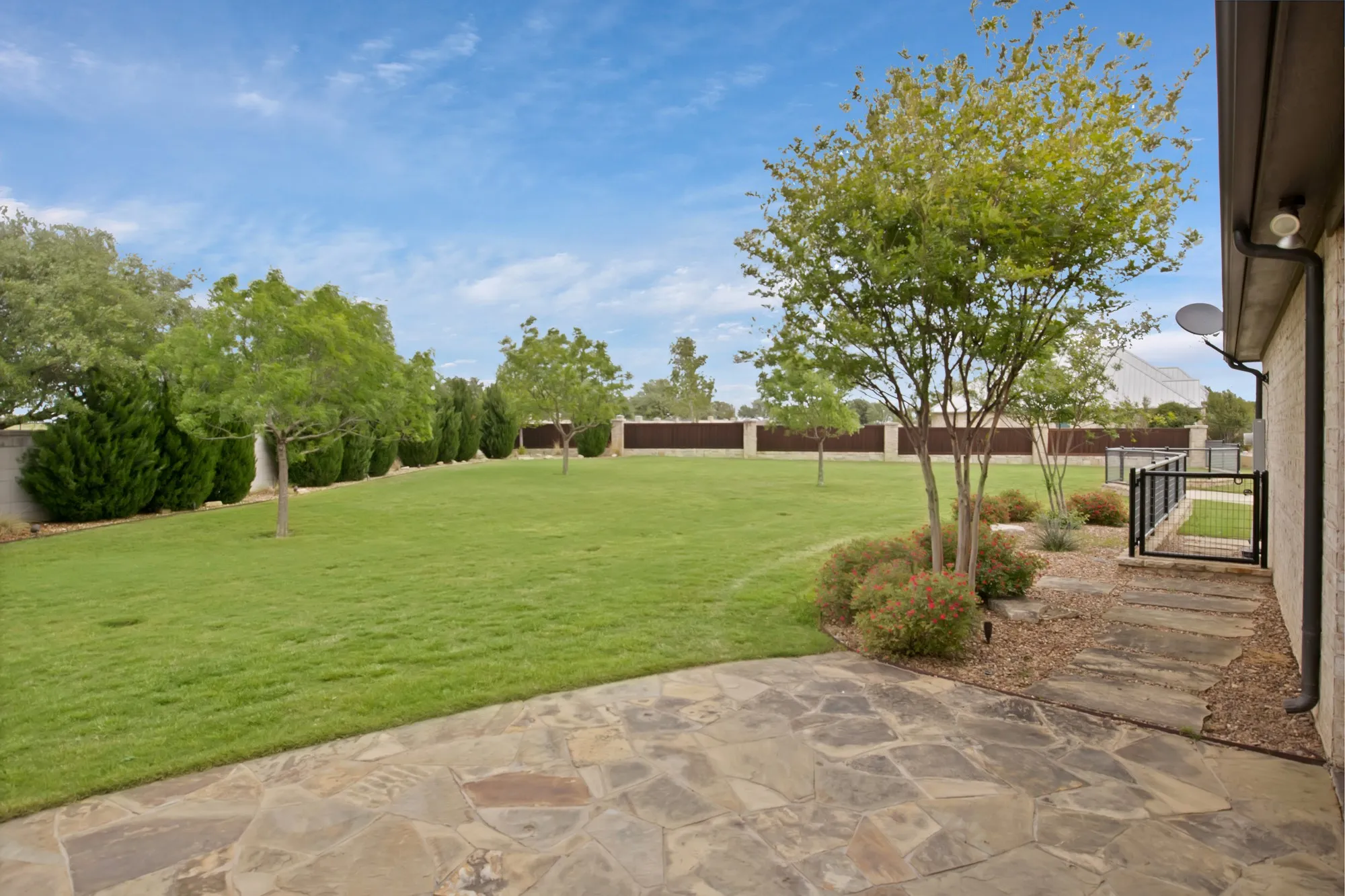 Expansive backyard featuring a natural stone patio, a large grass lawn, mature trees, ornamental shrubs, and a black wrought iron gate