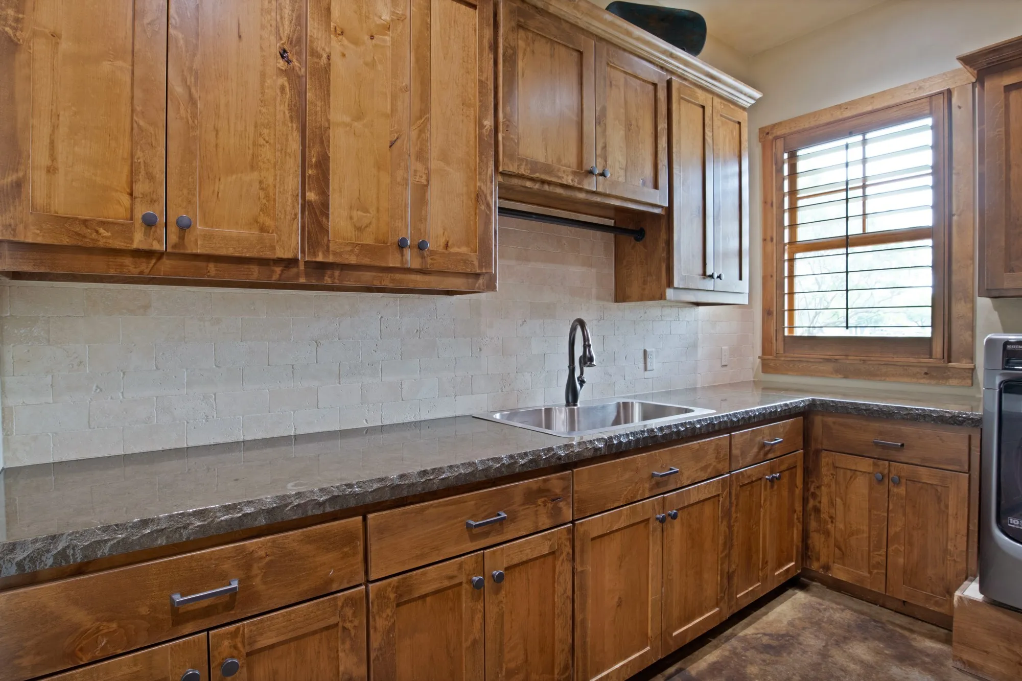 Wood cabinetry with dark hardware, a stainless steel sink with a gooseneck faucet, light-toned tiled backsplash, dark countertops, and a window with wood plantation shutters