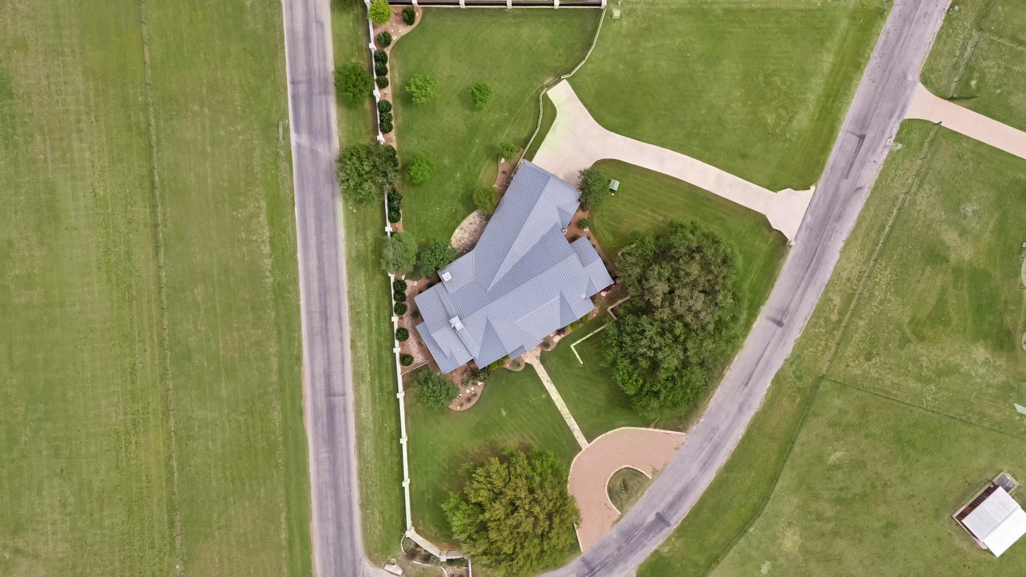 Expansive property featuring a grey multi-pitched roof, paved driveway, mature trees, manicured lawns, and a perimeter fence