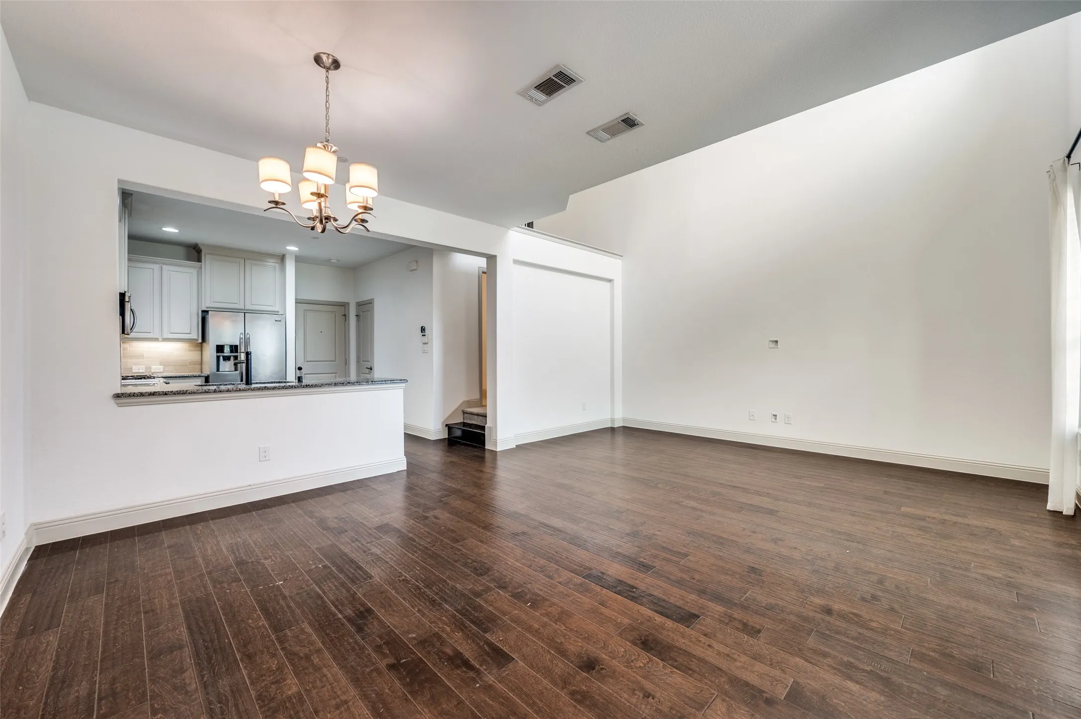 Large unfurnished living room with hanging lights, dark wood finished floors, and multiple windows bringing in natural light, showcasing flexible furniture layout options and open sightlines into adjacent spaces, ideal for homeowners who want a McKinney Texas townhouse with a big room for gatherings, entertaining, and daily living near major DFW employment centers.