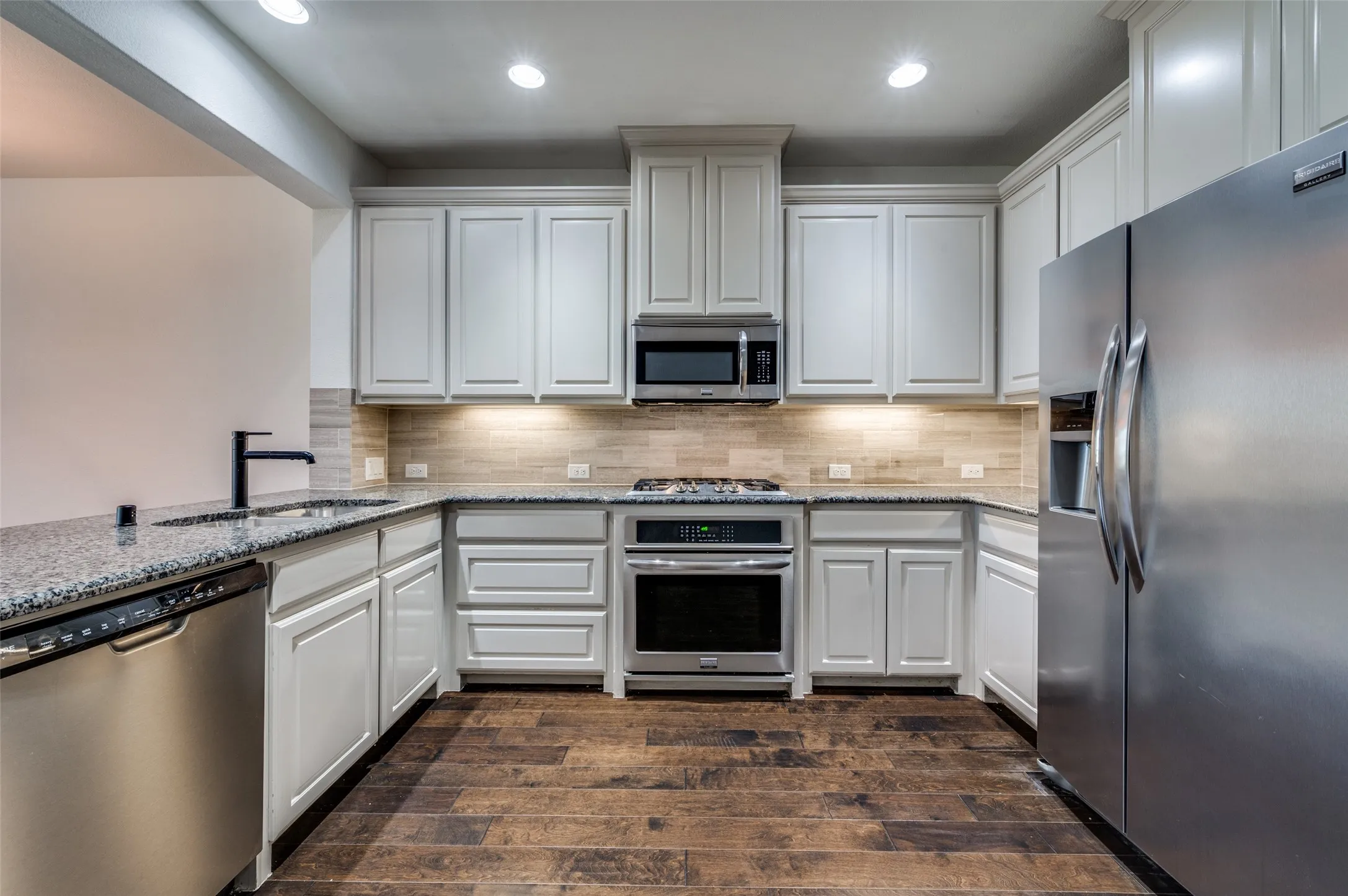 Gourmet style kitchen in McKinney Texas townhome featuring stainless steel appliances, light stone countertops, dark wood type flooring, white cabinets, recessed lighting, and tile backsplash, delivering the updated kitchen features and clean modern look today’s Collin County buyers search for when shopping online for McKinney TX townhomes for sale with upgraded kitchens.