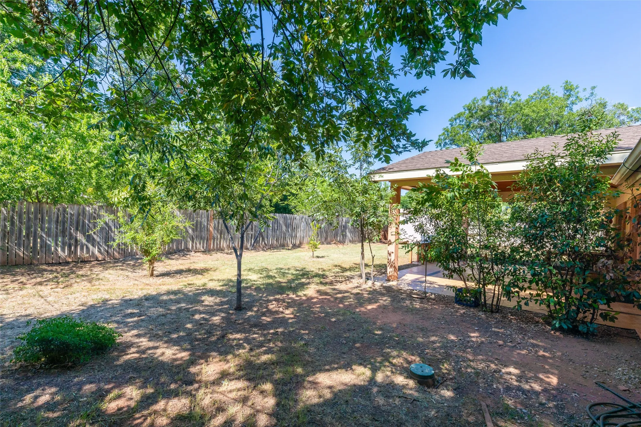 Spacious backyard featuring multiple mature trees providing natural shade, a full wooden privacy fence, and a covered patio area with a stone-clad exterior