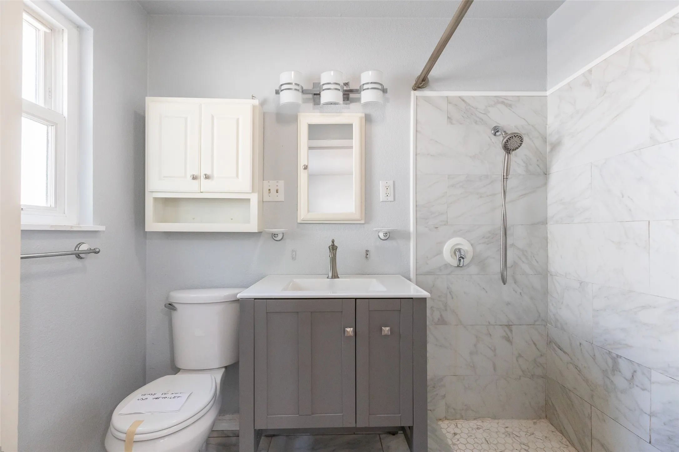 Bathroom featuring a gray vanity with an integrated sink, a white wall-mounted cabinet, a mirrored medicine cabinet, a tiled shower with a handheld showerhead, and a window providing natural light