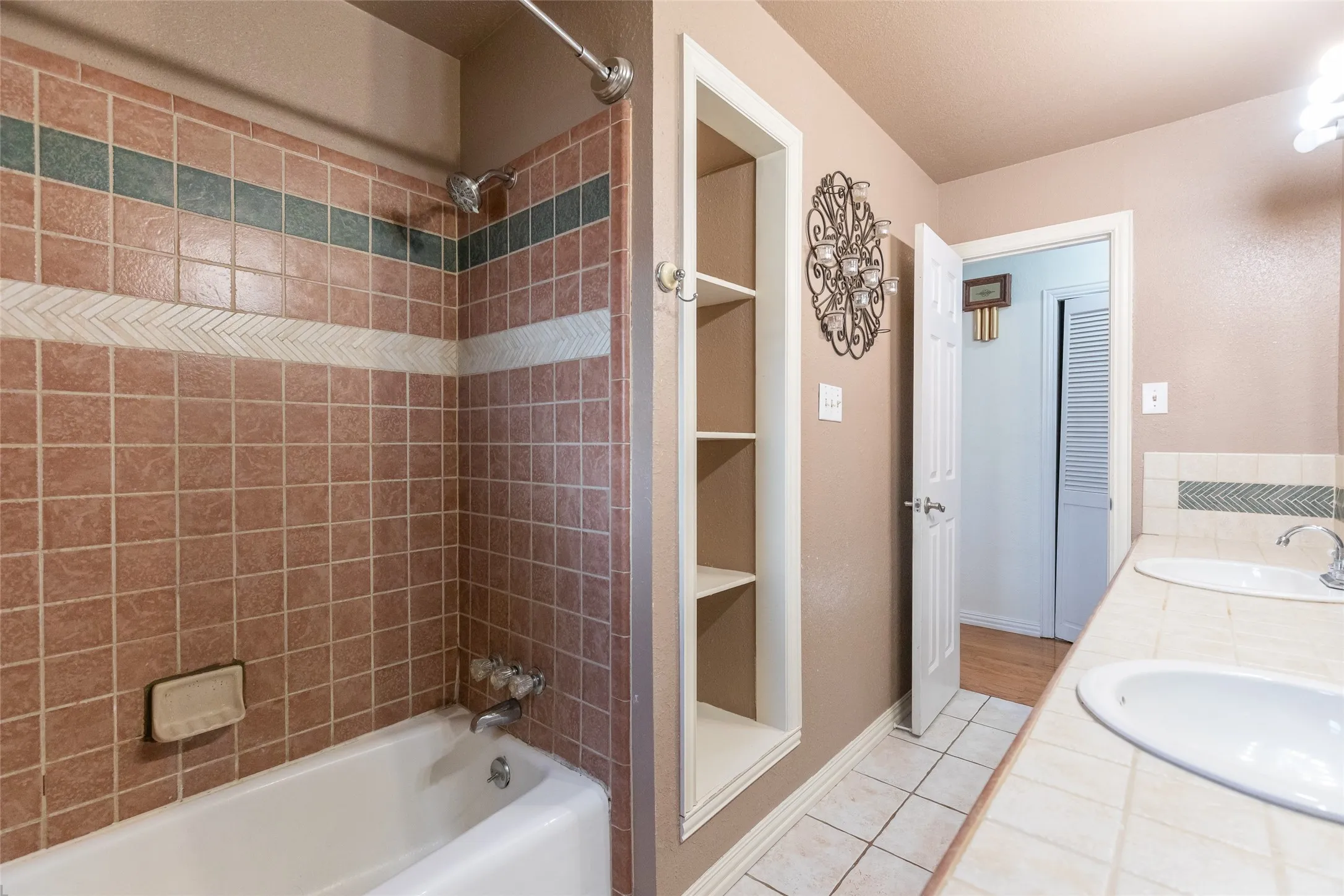 Bathroom featuring a dual-sink vanity with tile countertop, built-in shelving, tile flooring, a bathtub, and tiled shower surround with decorative accents