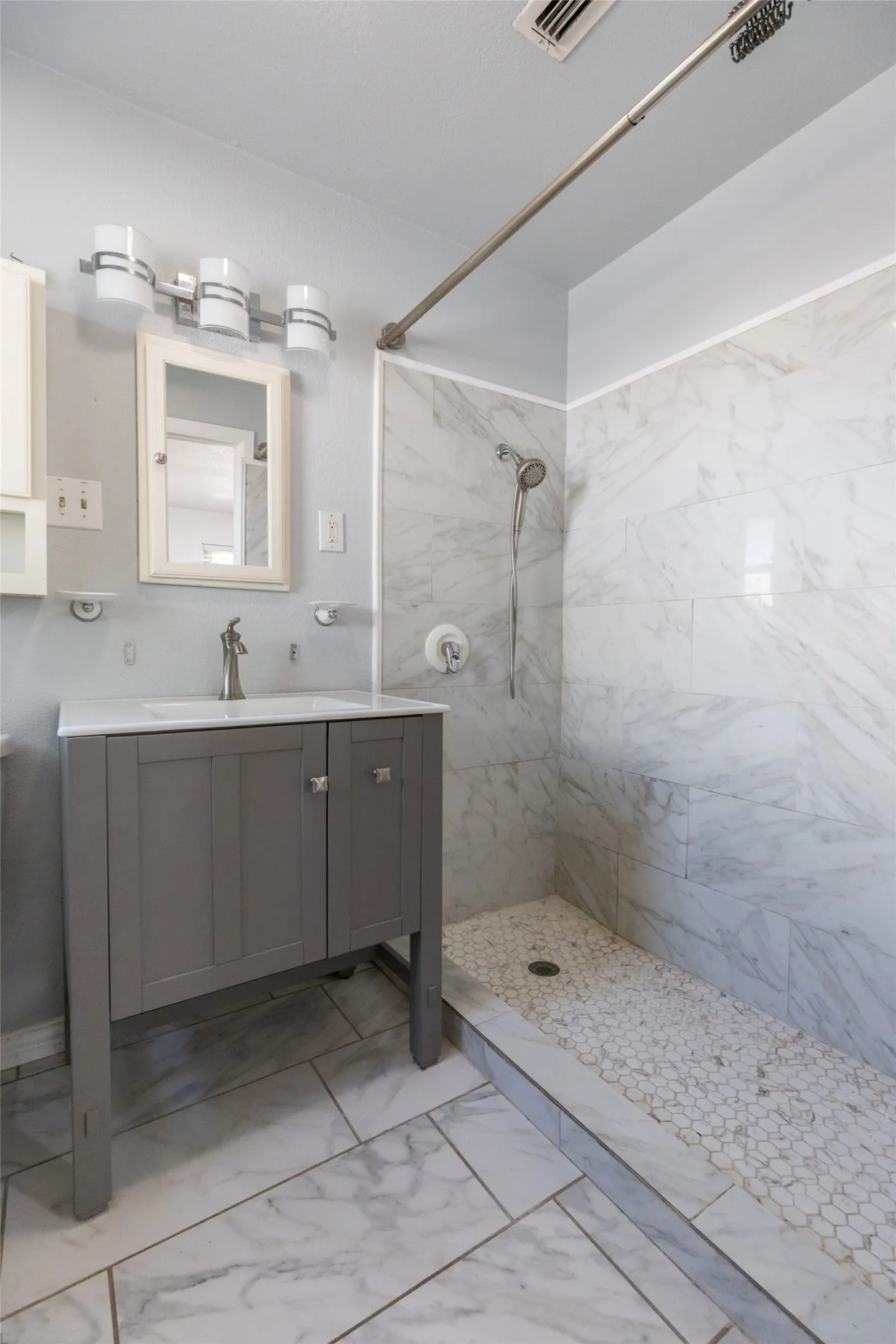 Bathroom featuring a walk-in shower with large format tile walls and a pebble floor, a single vanity with a gray cabinet and white countertop, and rectangular floor tiles
