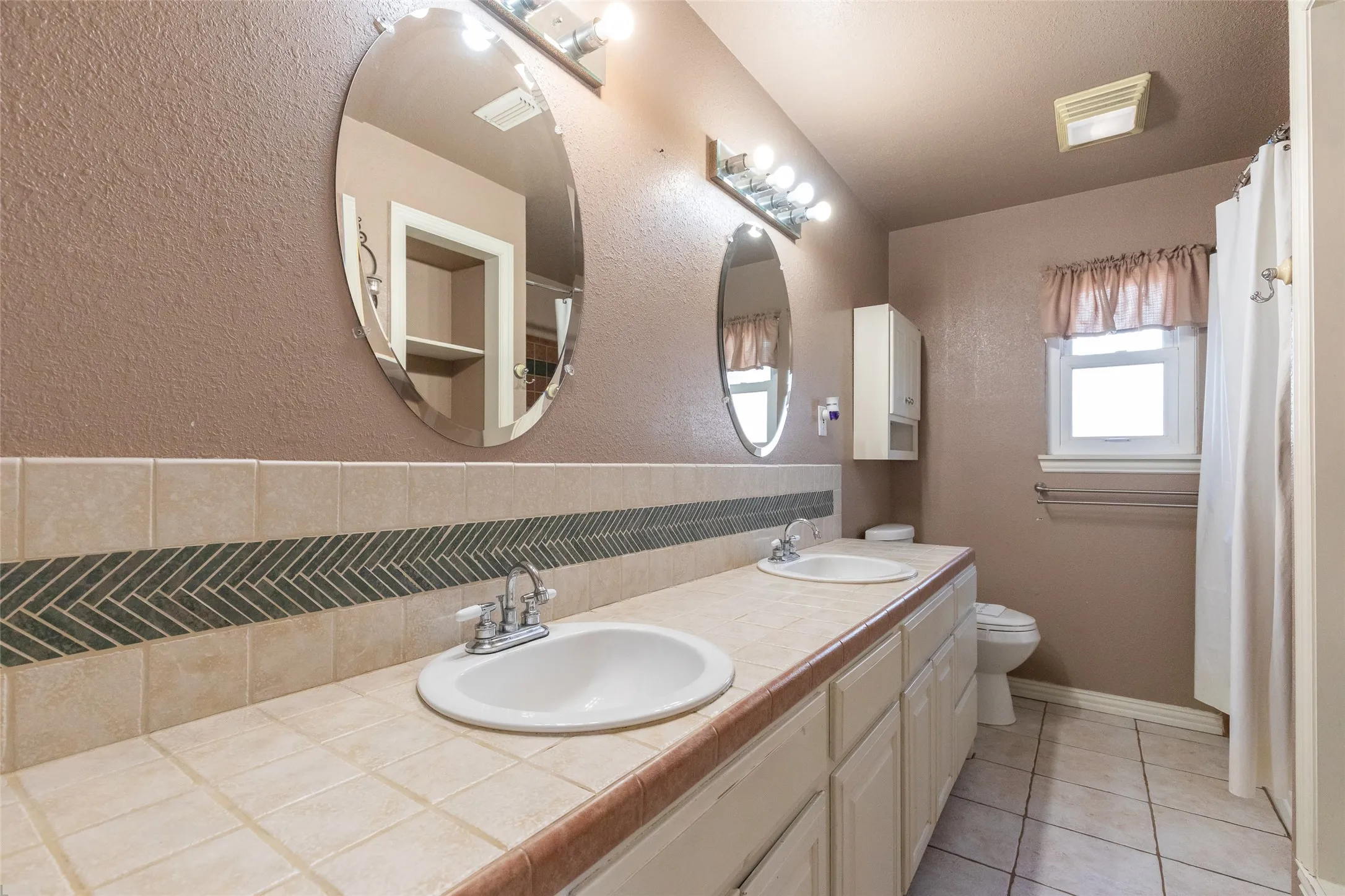 Bathroom featuring a double vanity with integrated sinks, a tiled countertop, and a decorative herringbone tile backsplash