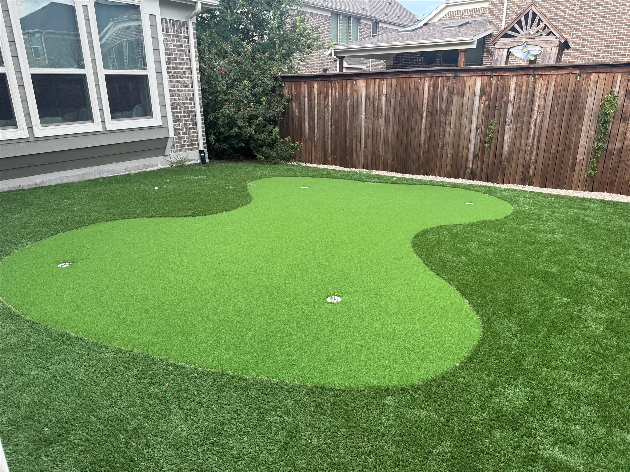 Manicured artificial turf putting green with multiple cups, bordered by natural grass and a wood privacy fence