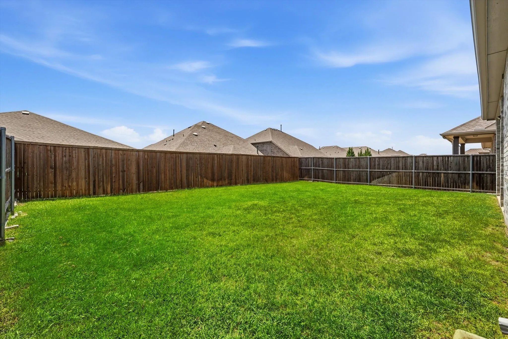 Expansive backyard featuring a large green lawn, full wood privacy fencing, and a partial view of an exterior brick wall with an attached covered patio