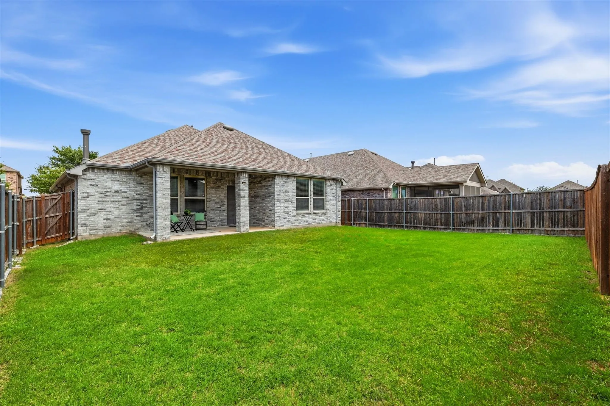 Expansive backyard showcasing a pristine manicured lawn with mature landscaping, fenced perimeter, and a single-story stone exterior home featuring a pitched roof, multiple windows, and an attached chimney