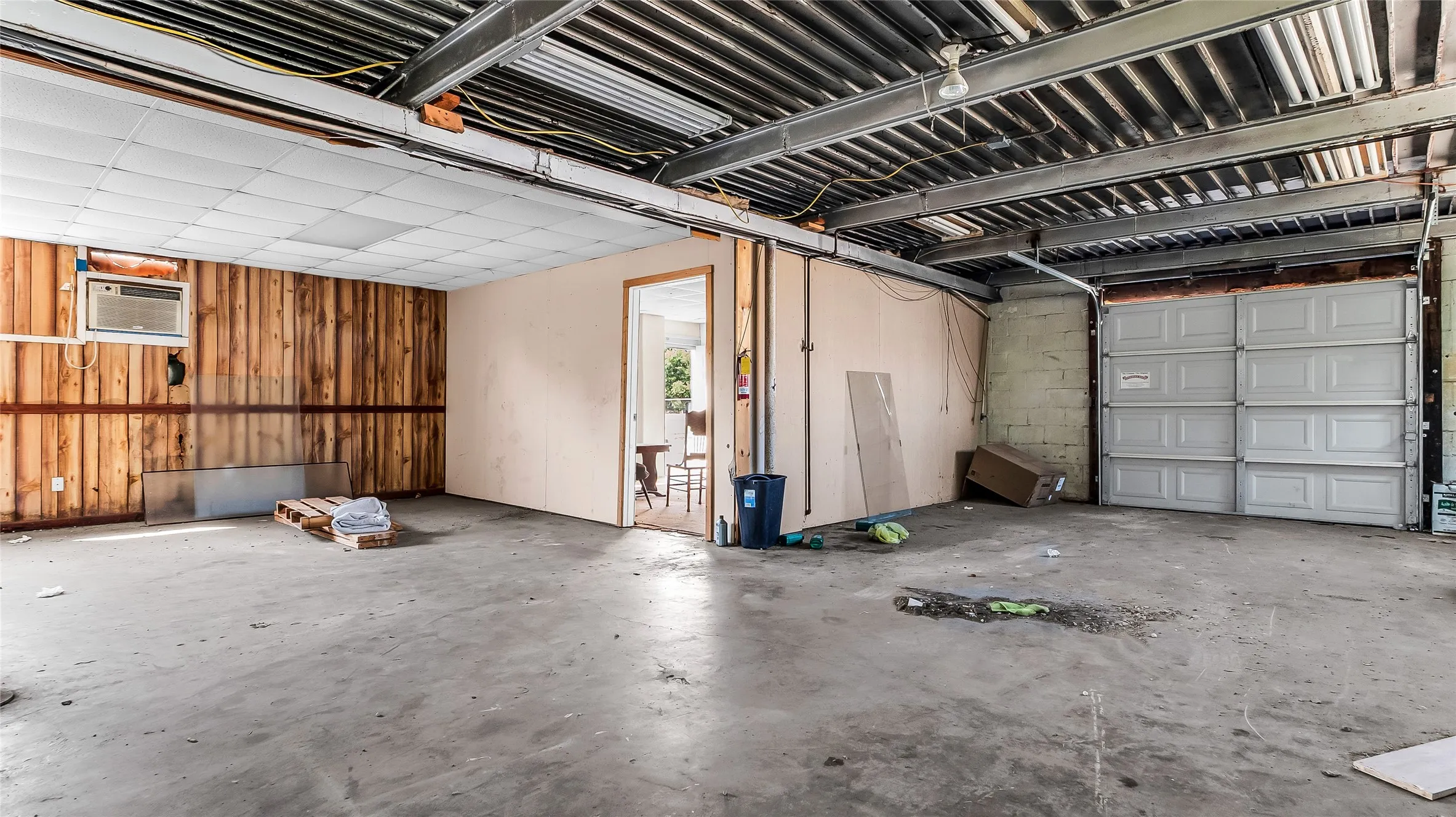 Concrete flooring, exposed ceiling joists, wood paneling, paneled garage door, and a wall-mounted air conditioning unit