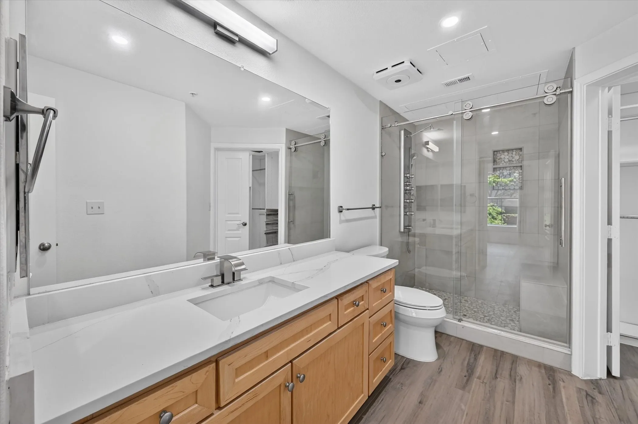 Bathroom featuring a light wood-finish vanity with a white countertop and undermount sink, a large mirror with integrated lighting, a glass-enclosed shower with pebble flooring and a multi-function shower panel, and wood-finish flooring