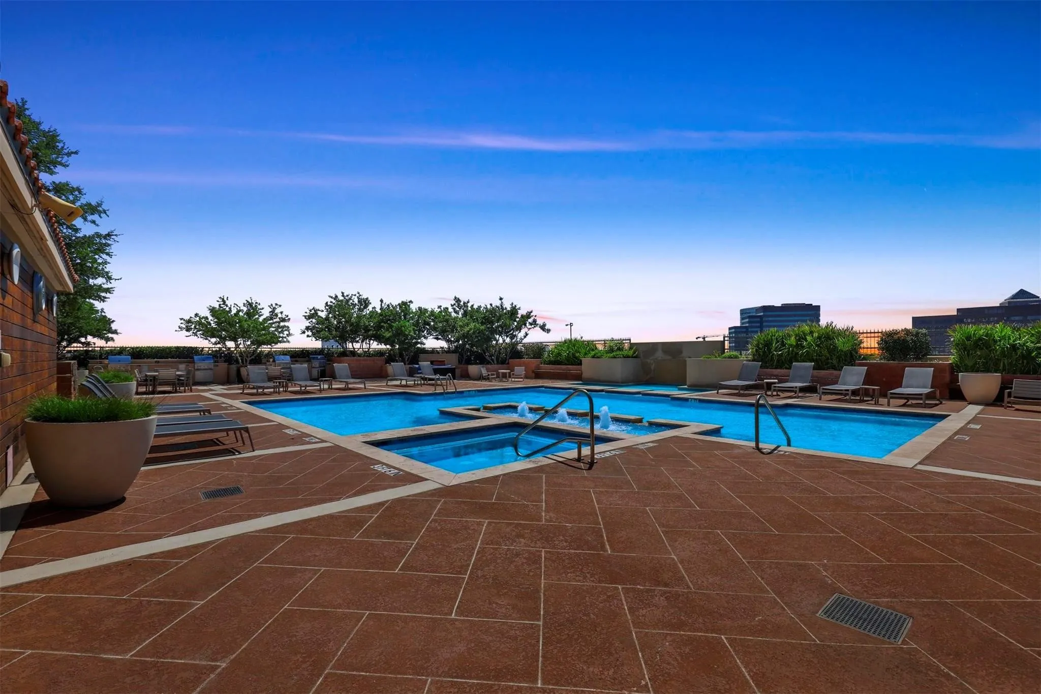 Resort-style rooftop pool deck featuring a sparkling blue swimming pool with integrated spa, expansive terracotta-toned paver decking, mature landscaping with specimen trees, and panoramic city skyline backdrop