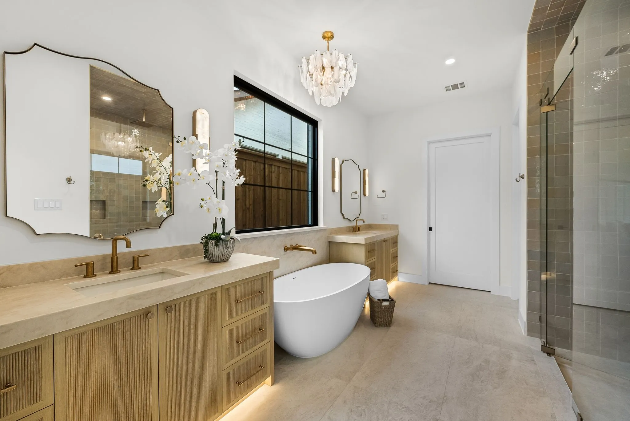 Spacious bath featuring dual wood-finish vanities with light-toned countertops, a freestanding soaking tub, a large walk-in shower with glass enclosure, and a statement chandelier