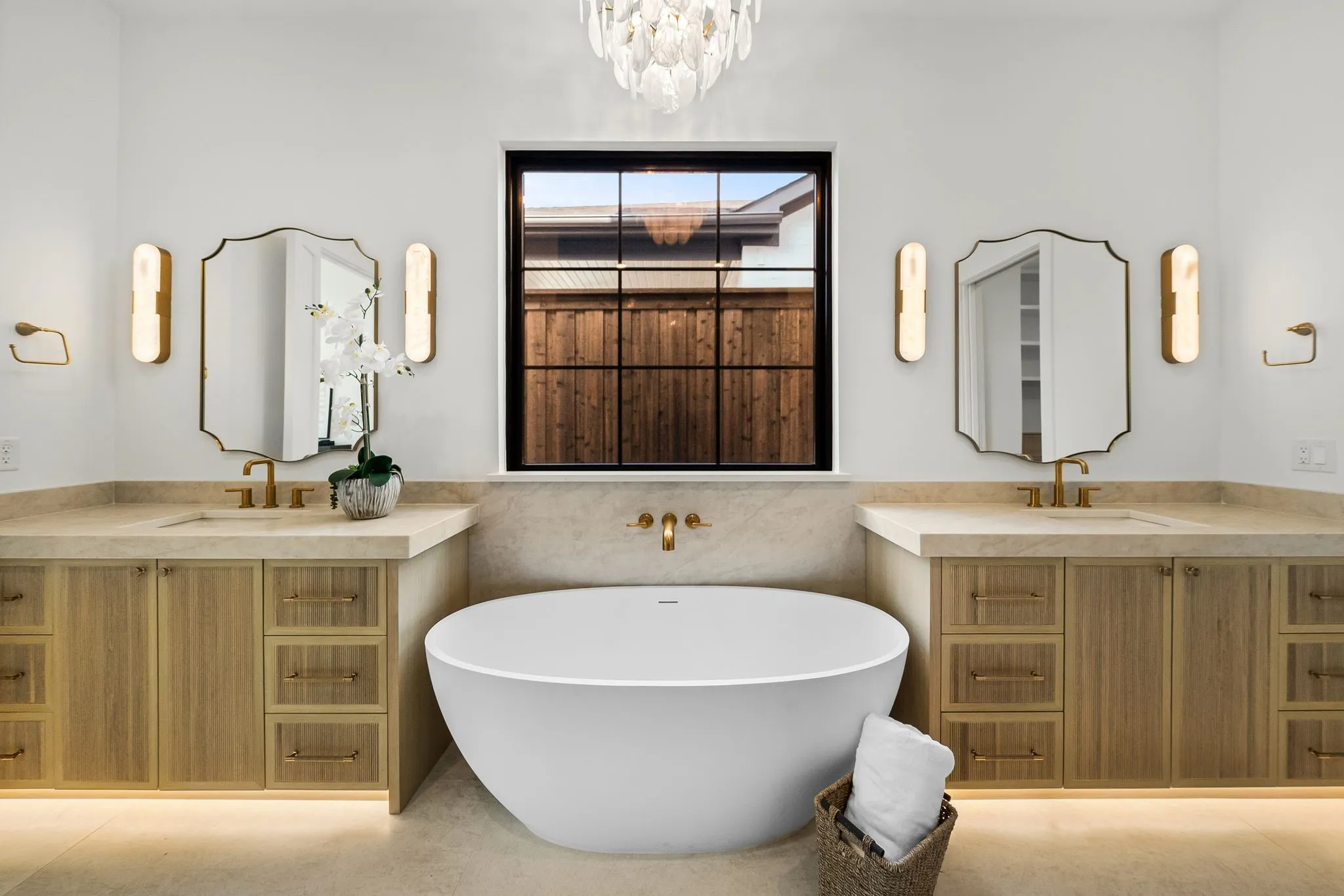 Spa-inspired primary bathroom featuring dual wood-finish vanities with integrated storage, dual framed mirrors with integrated sconce lighting, a freestanding soaking tub, heated flooring, and a black-framed window with interior shutters overlooking the landscape