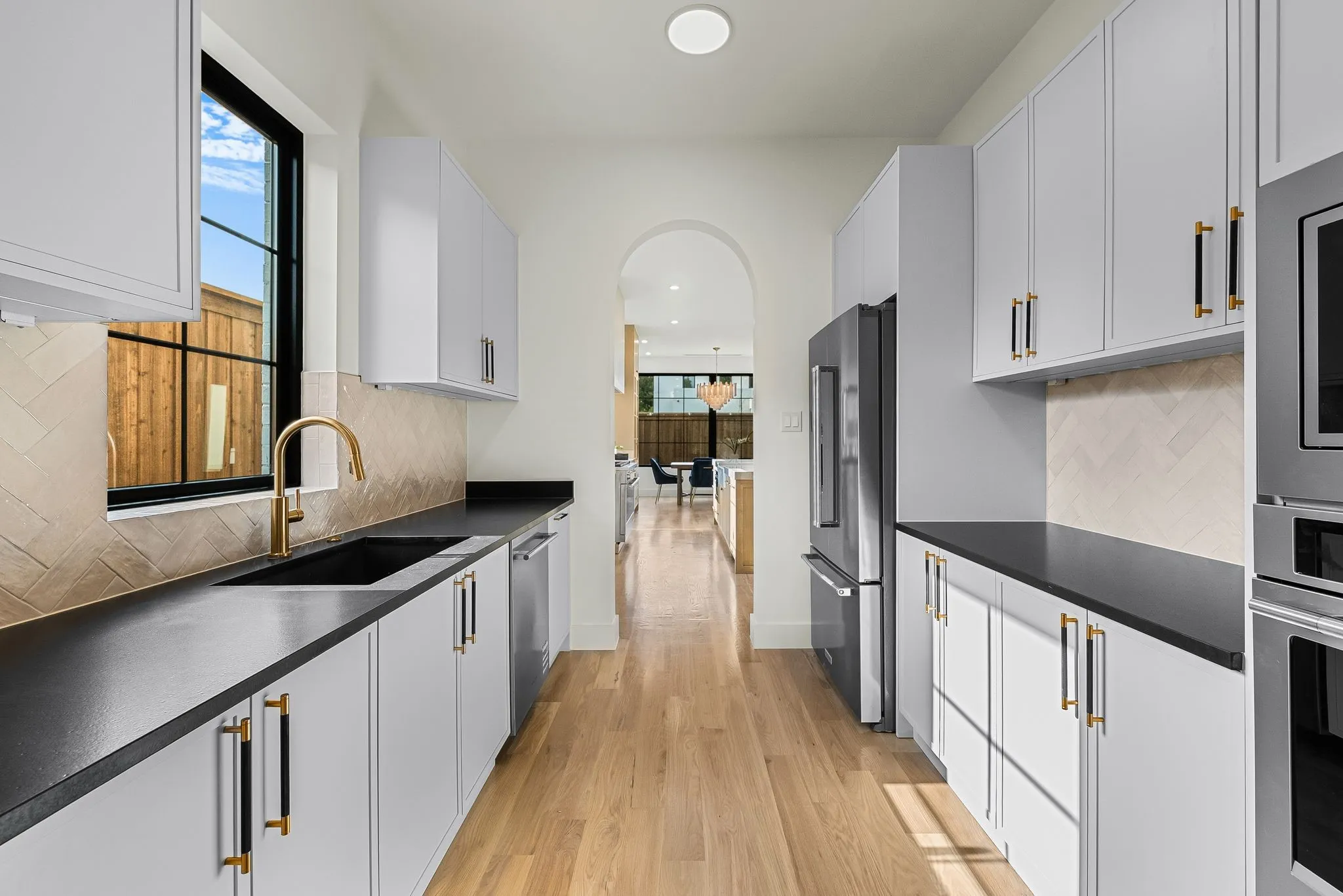 Modern galley kitchen featuring light wood-finish flooring, white cabinetry with contrasting hardware, dark countertops, a herringbone tile backsplash, and a black-framed window
